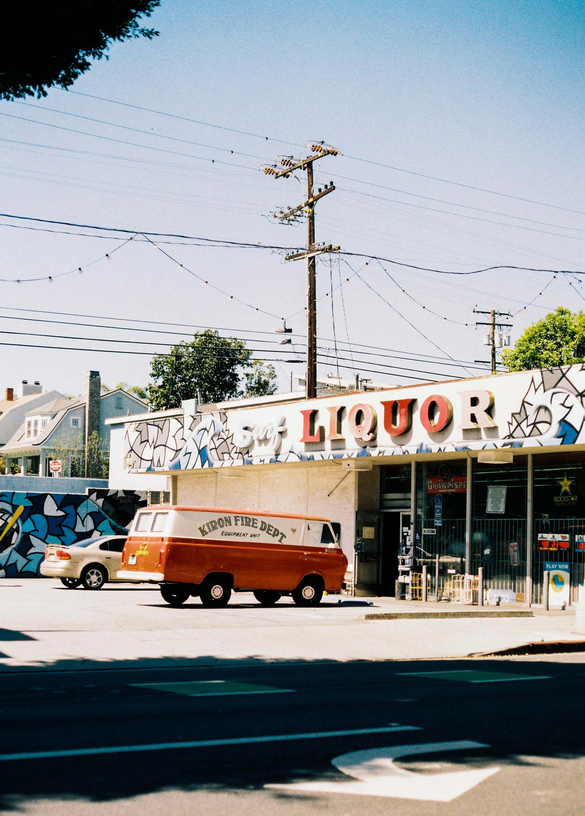 A storefront with a sign that says 'Liquor' in large red letters, a mural with geometric patterns on the wall, a vintage orange and cream fire department vehicle parked outside, and trees and houses in the background.