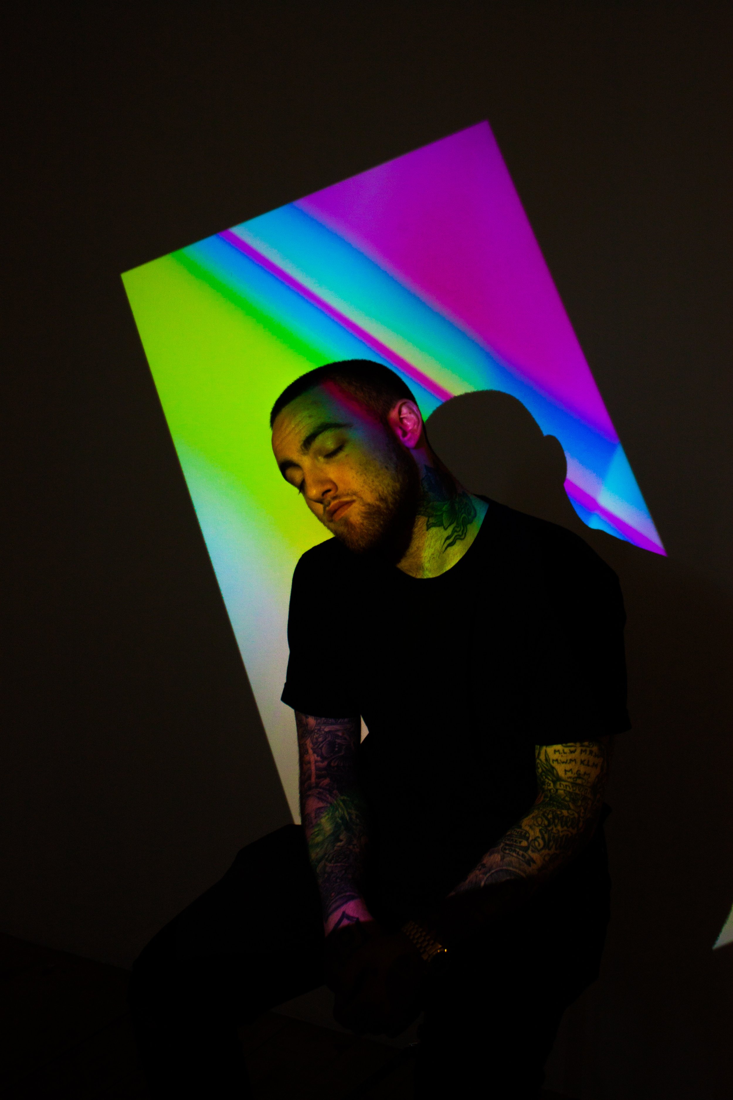 A man with tattoos on his arms and neck, wearing a black shirt, sitting with eyes closed against a dark background illuminated by a colorful, geometric light projection.