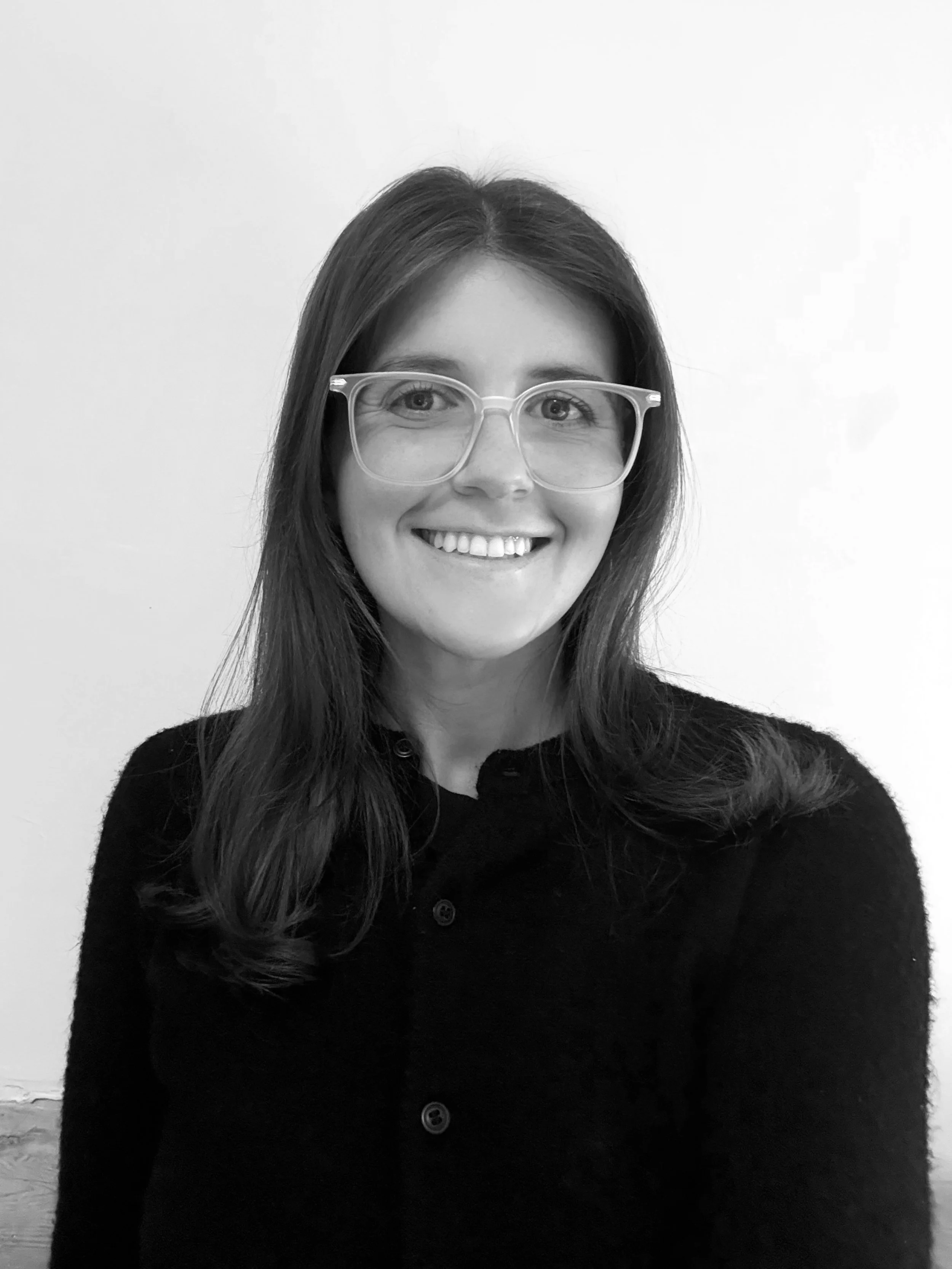 Black and white photo of a woman with long hair, glasses, and a smile, wearing a dark button-up sweater, against a plain light background.
