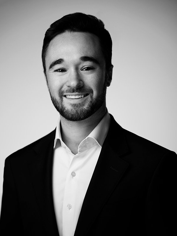 Black and white portrait of a young man with dark hair and beard, smiling, wearing a suit jacket and white dress shirt against plain background.