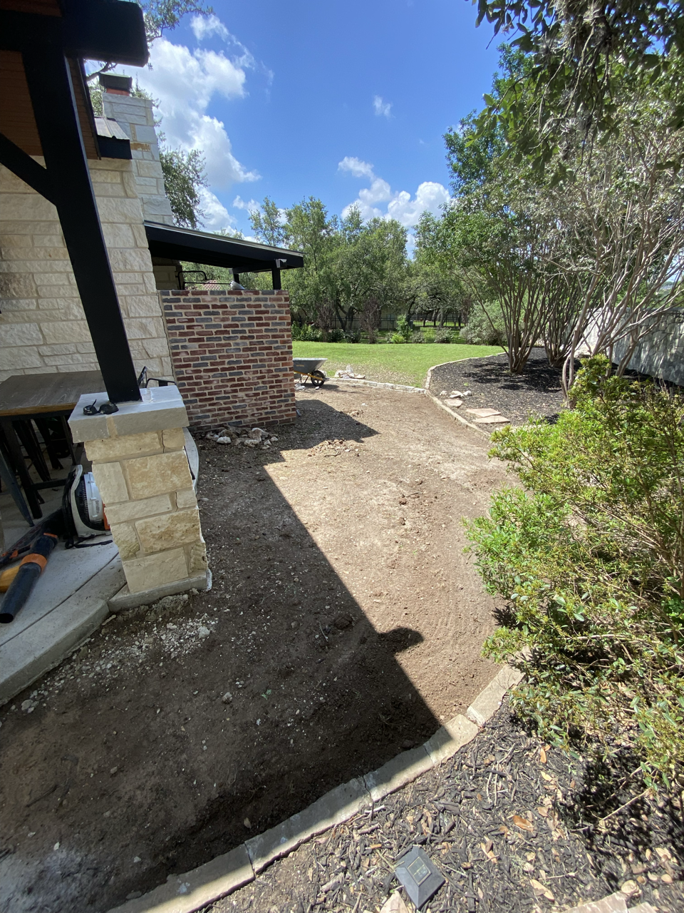 Backyard with dirt area, surrounded by bushes and trees, under a bright blue sky with clouds, part of a house with brick and stone exterior, construction materials and tools nearby.