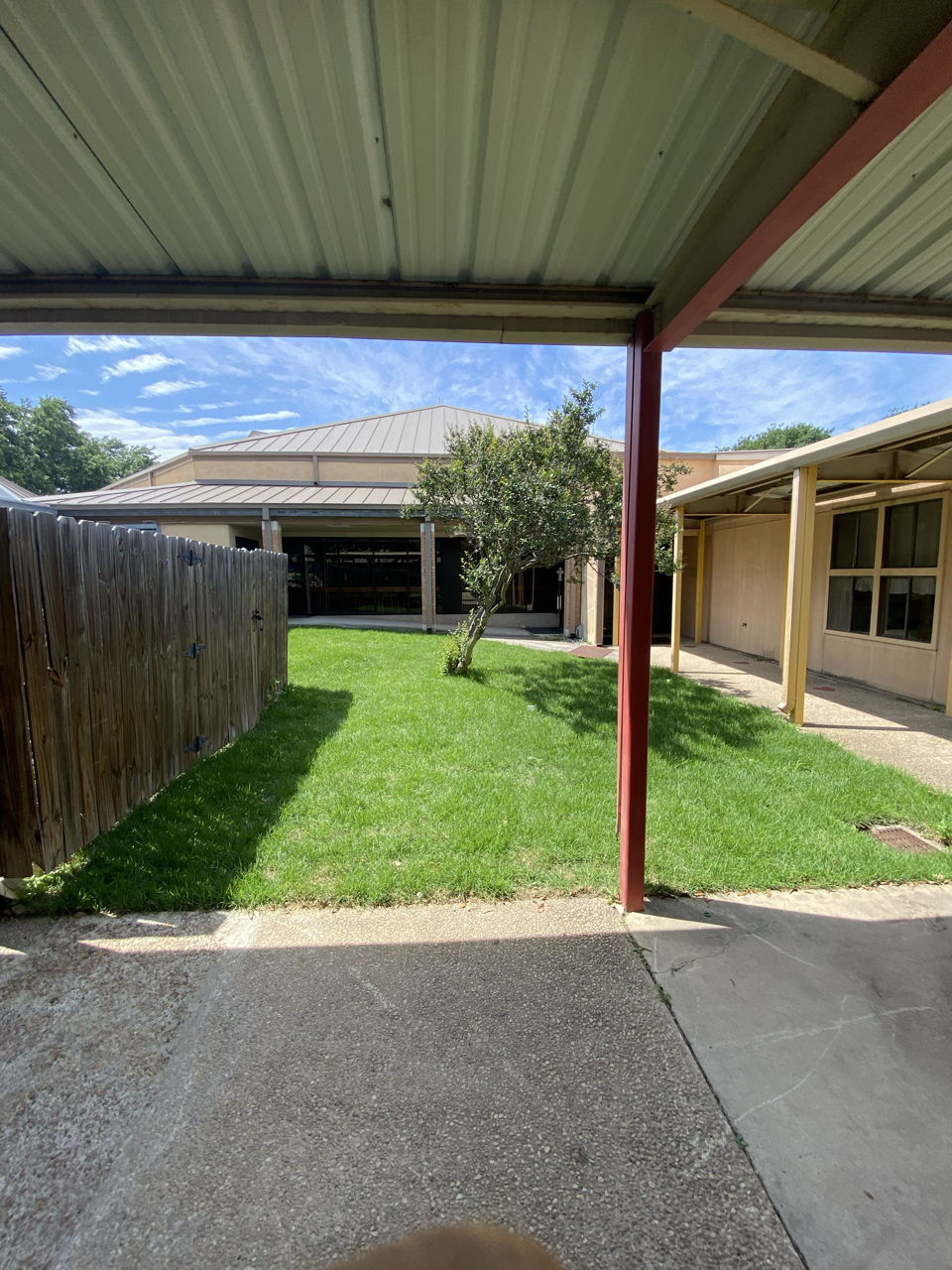 A backyard view from under a carport, showing a grassy lawn with a small tree, a wooden fence on the left, and neighboring buildings with roofs, windows, and support beams in the background under a partly cloudy sky.