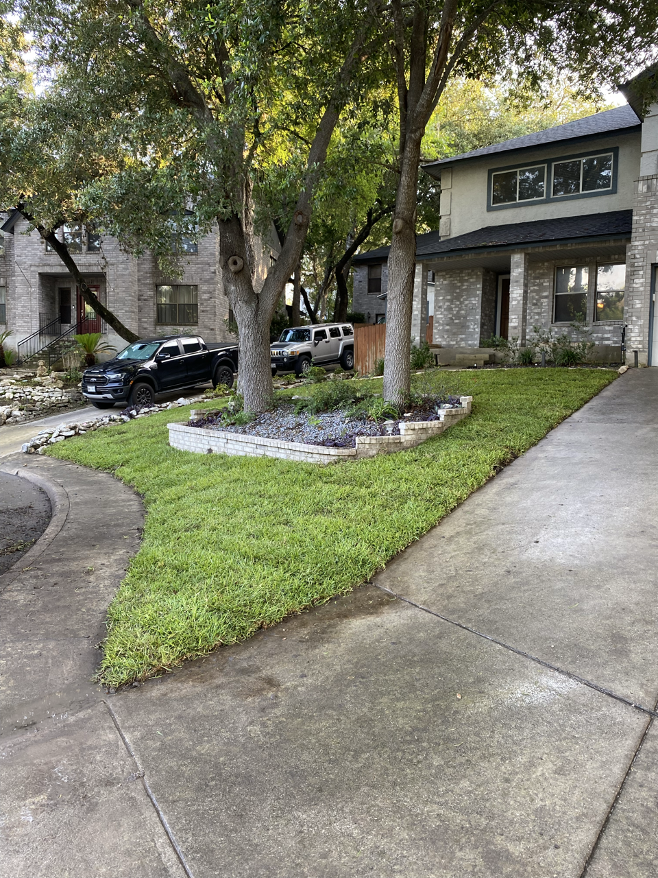 View of a residential driveway with a grassy lawn, trees, and houses in the background. Two black vehicles are parked along the street. The driveway is wet, and the house is made of brick with a dark roof.