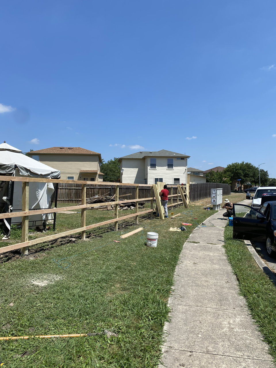 People constructing a wooden fence along a sidewalk in a residential neighborhood, with houses and parked cars in the background under a clear blue sky.
