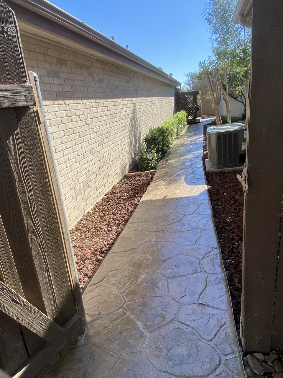 Side yard pathway along a brick house with bushes, trees, a lattice structure, an air conditioning unit, and clear blue sky.