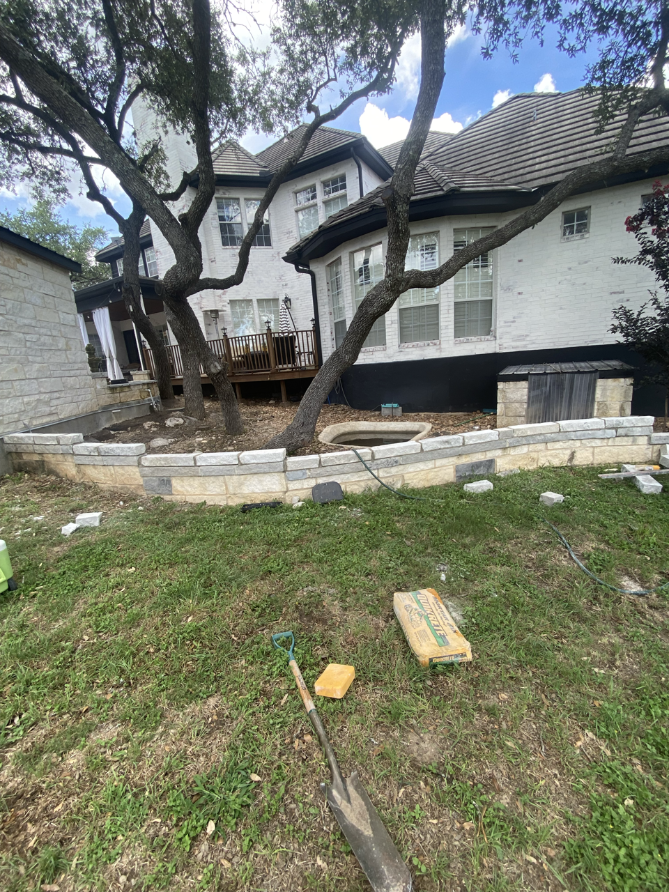 Backyard with trees and a brick retaining wall under construction, tools and materials on the grass, and a white house with large windows and a wooden deck in the background.
