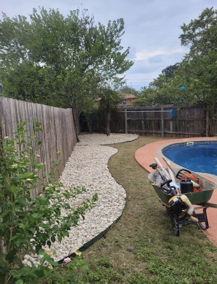 Backyard with a curved white stone pathway, wooden fence, green trees, a pool with a red border, and a wheelbarrow with yard tools.