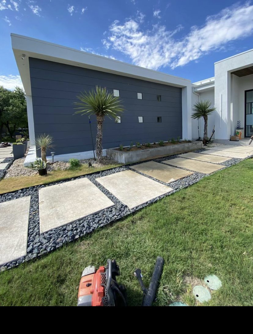 Modern house with a dark gray garage door, landscaped yard with concrete stepping stones and black rock borders, three palm trees, and side patio area under blue sky.