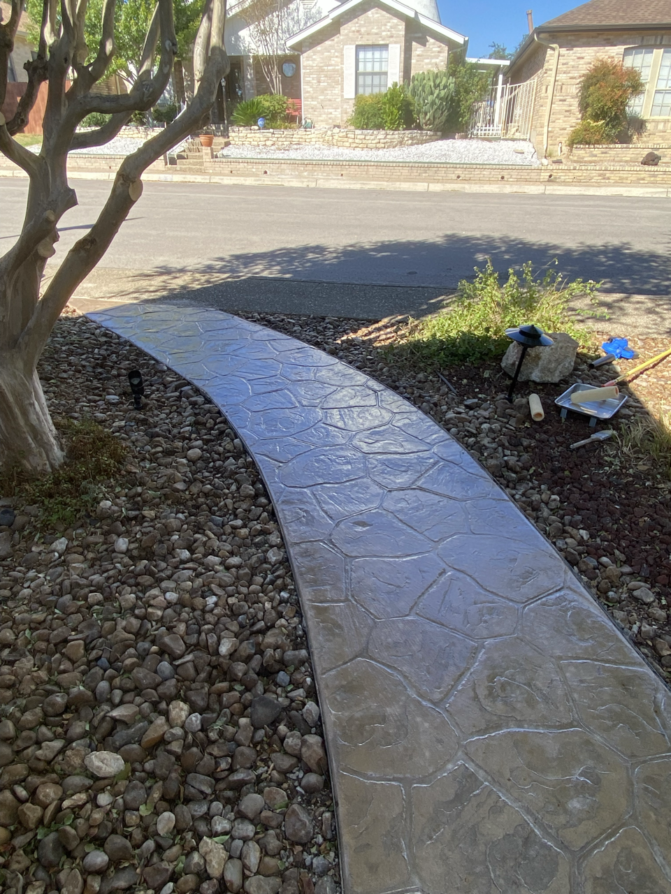 Newly paved concrete sidewalk in front of a house, surrounded by rocks and garden decor, with trees and neighboring houses visible in the background.