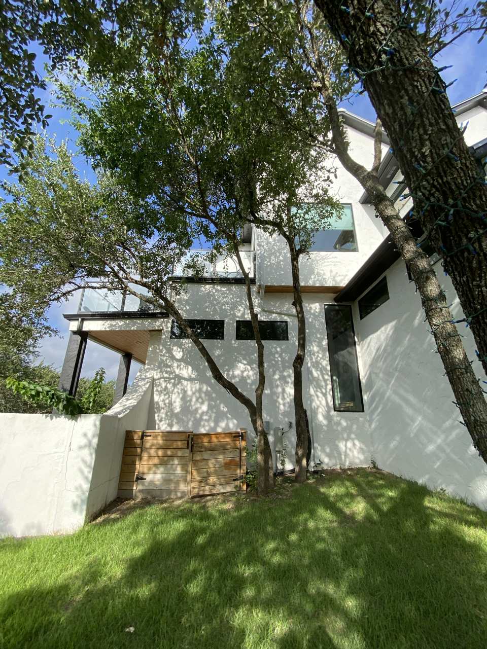 Modern white house with large windows, surrounded by green lawn and trees, with sunlight casting shadows.