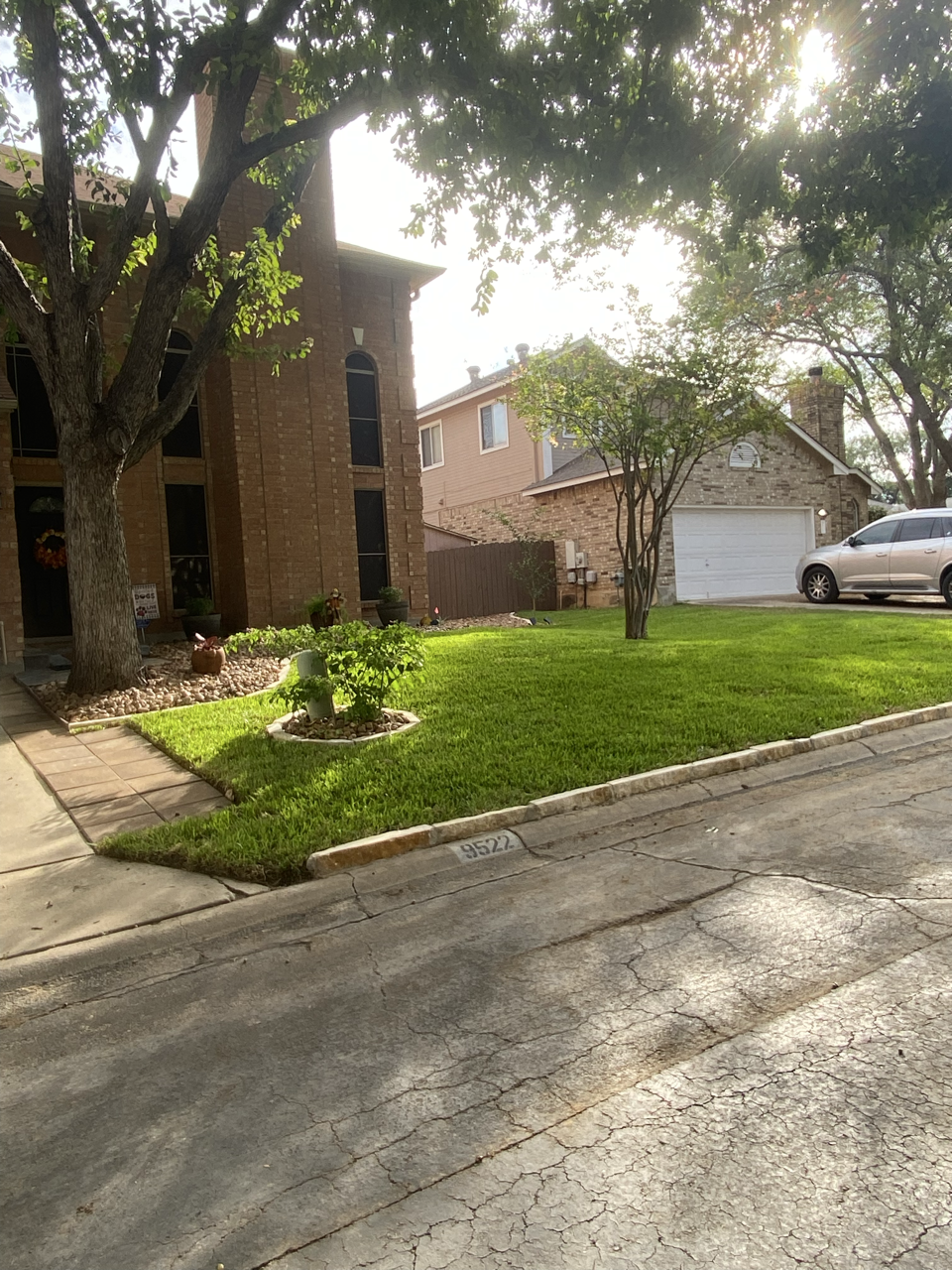 A residential driveway with a parked car next to a lawn with trees, garden decorations, and a house in the background.