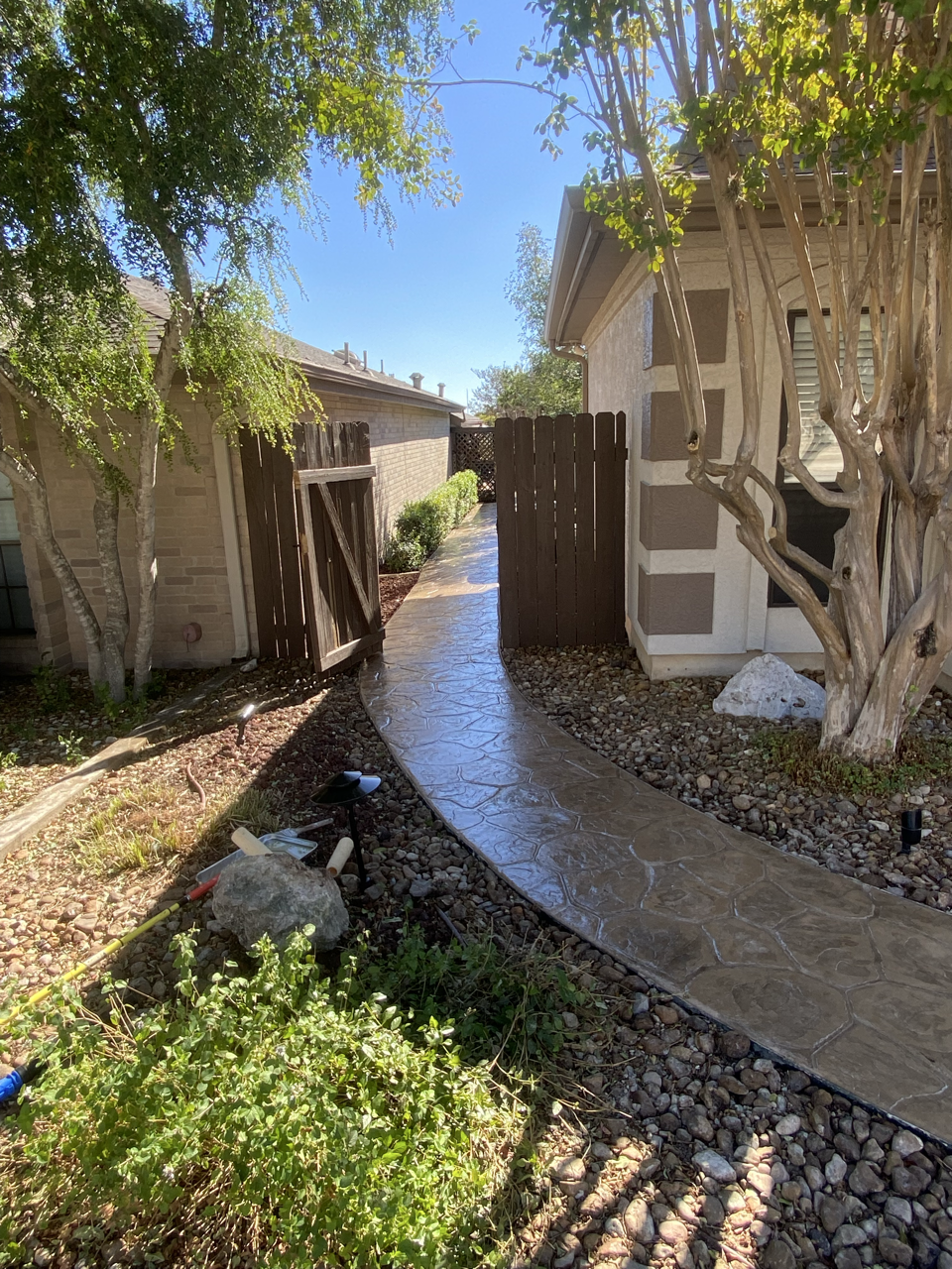 A narrow concrete walkway with stamped design, leading along a house with a beige exterior, next to a tall wooden fence and a small garden area with trees, bushes, and rocks, under a bright blue sky.