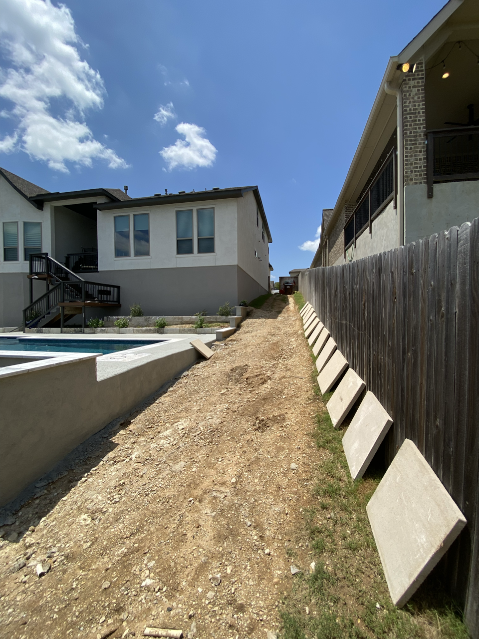 A dirt pathway running alongside houses on a sunny day, with a fence on the right side and a back yard with a pool on the left.