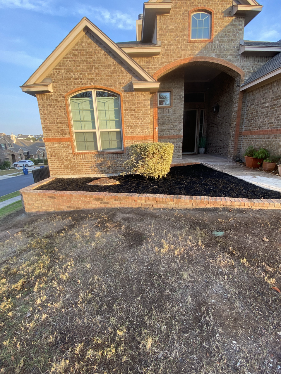 Front yard of a brick house with a flower bed, shrub, potted plants, and a walkway leading to the front door, under clear blue skies.