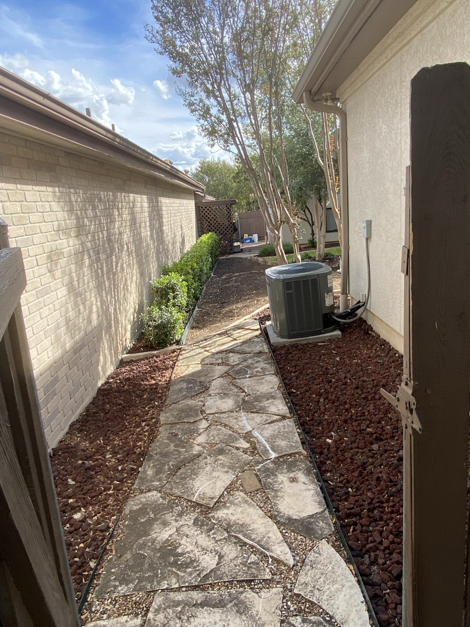 Backyard pathway with flagstone stones, bordered by mulch and greenery, with trees and a house wall, under a partly cloudy sky.