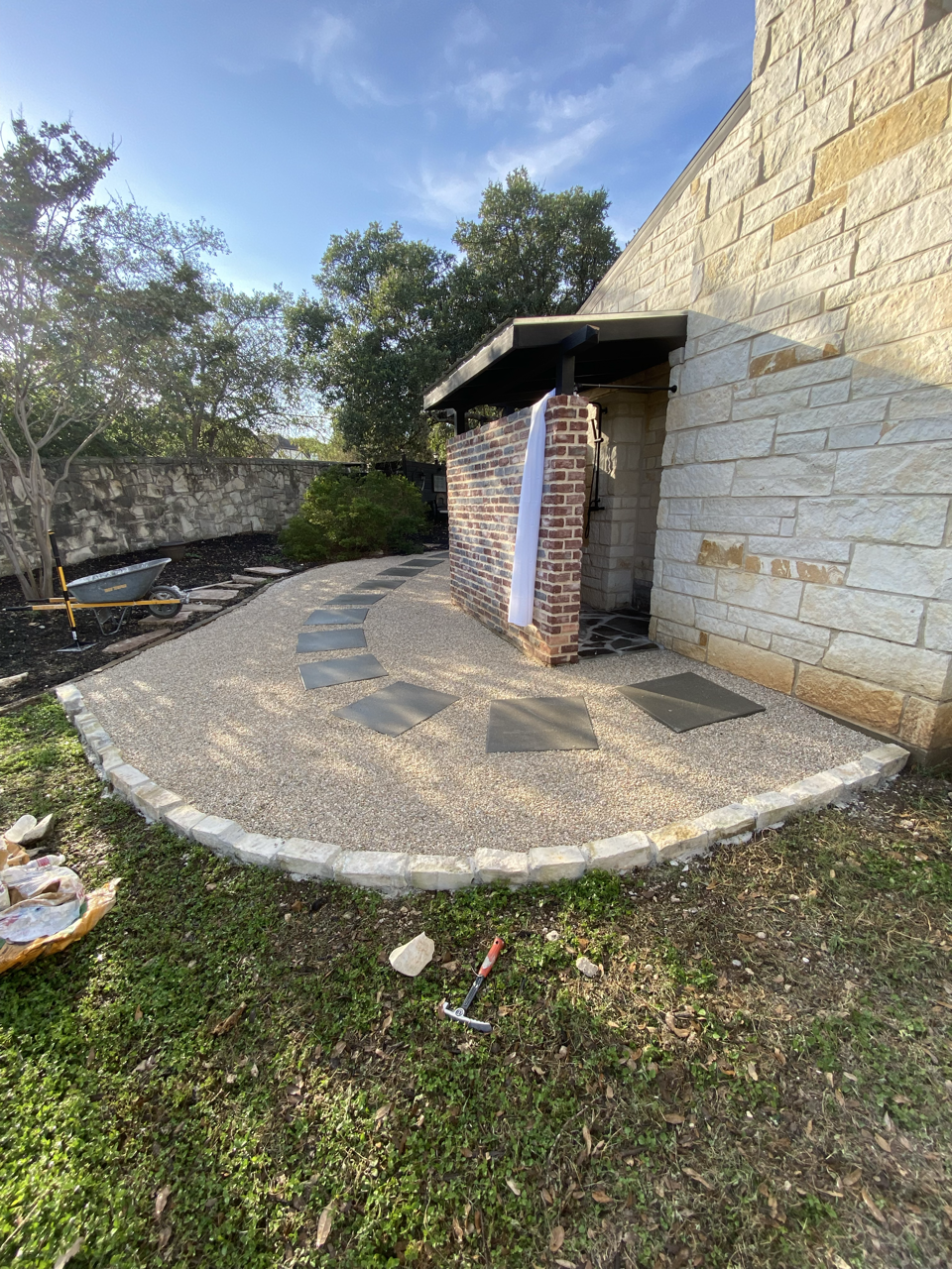 Backyard patio under construction with a curved pathway, stepping stones, brick and stone wall, and a small covered outdoor area.