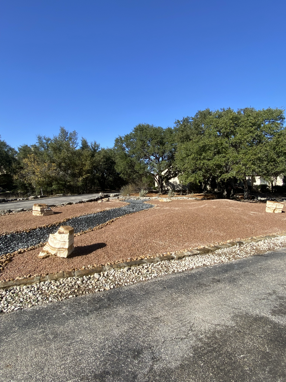 A landscaped area with gravel and rocks, bordered by a paved surface, with trees and a blue sky in the background.