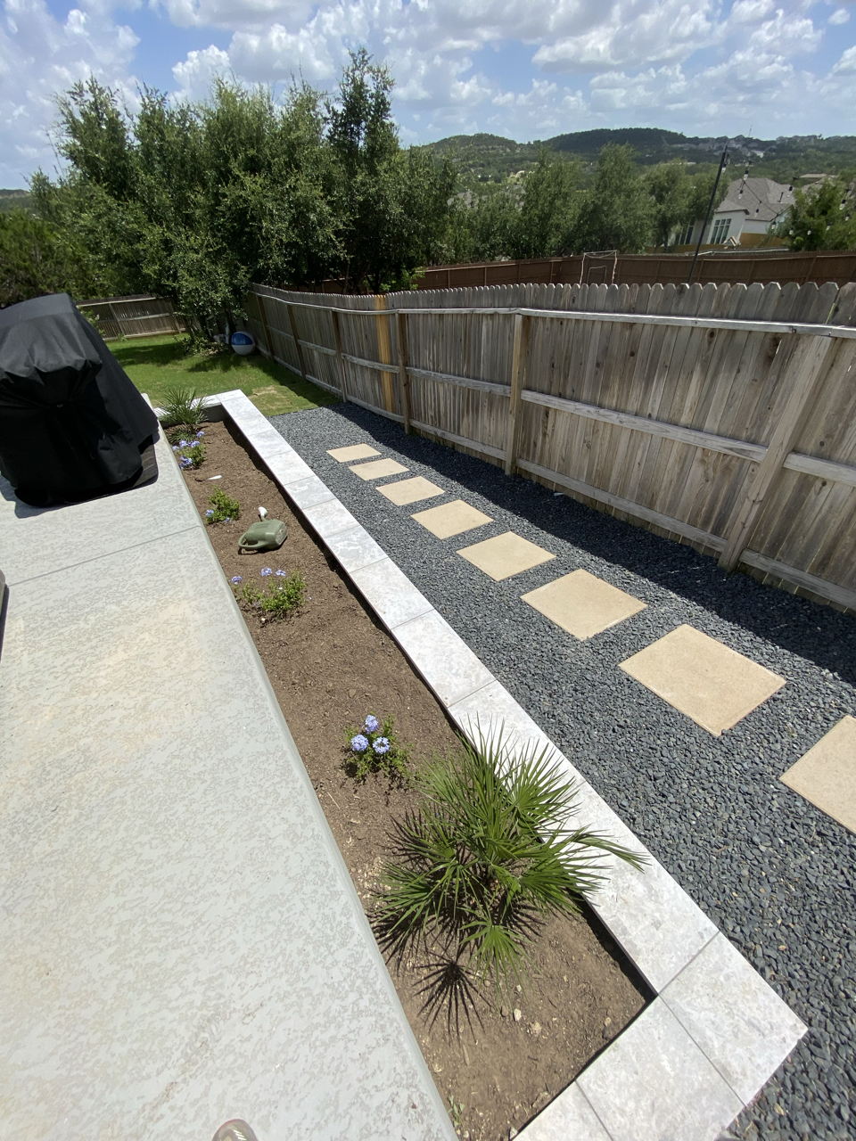Backyard with a concrete patio, small flower bed with plants, stepping stones on gravel pathway, wooden fence, sky with clouds, trees and hills in the background.