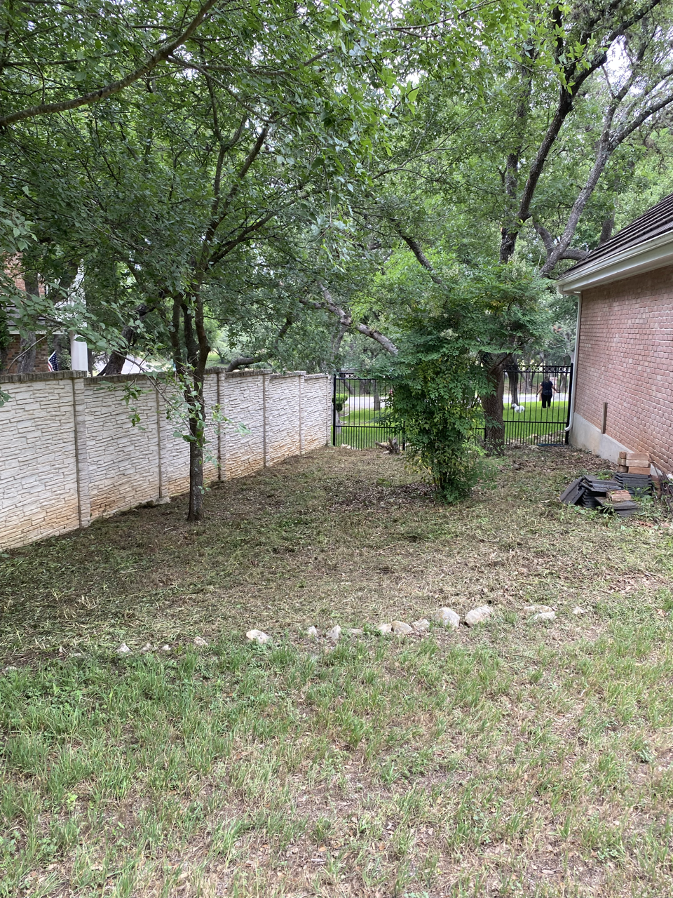 Backyard with green grass, trees, a stone fence on the left, a black metal gate at the center, and a brick house on the right.