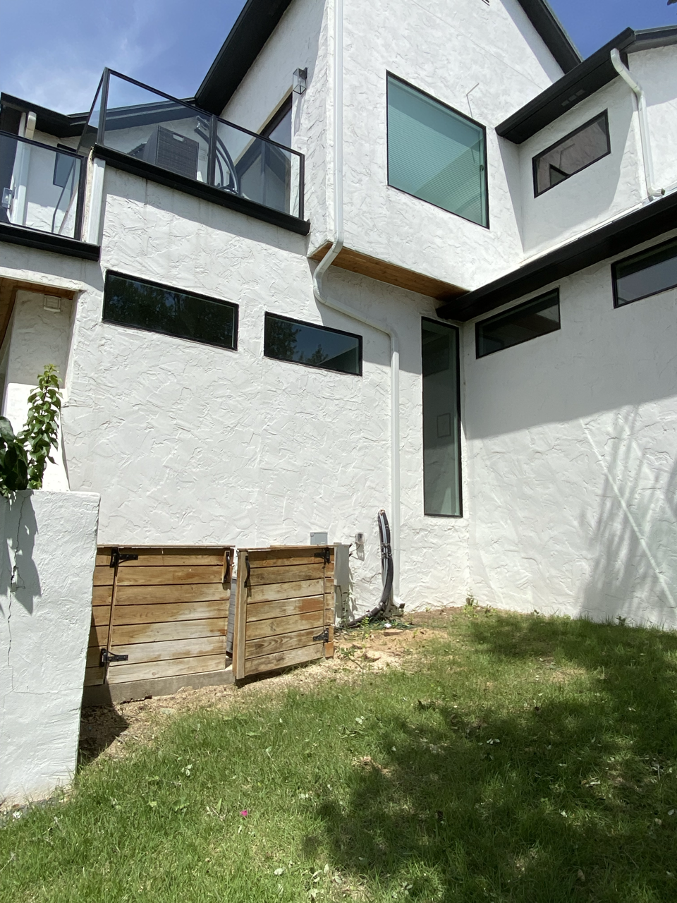 Backyard view of a modern white house with multiple rectangular windows, a small balcony with glass railing, and a wooden yard gate resembling a small fence. Green grass and small plants are in the yard.