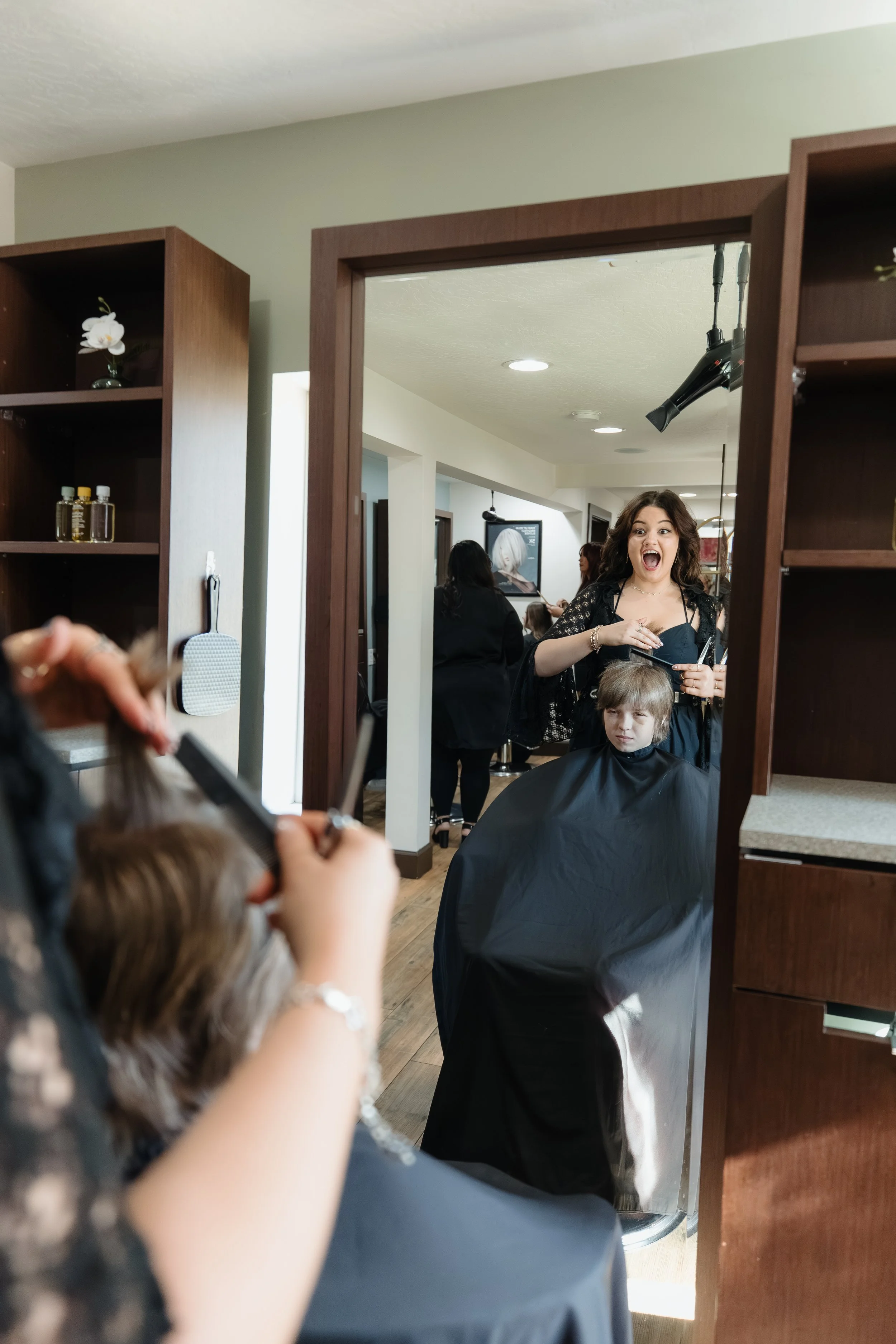 A woman at a hair salon giving a lively expression as she styles a young girl's hair who is sitting in a chair covered with a black cape, while another person is cutting the girl's hair.