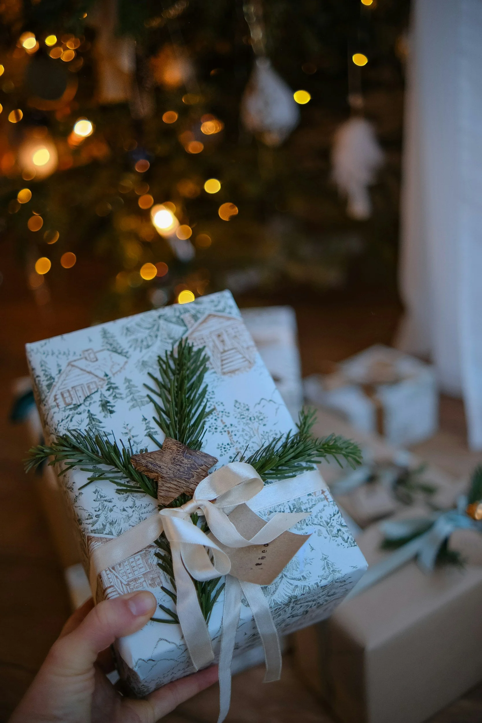 A person holding a wrapped Christmas gift decorated with a sprig of evergreen, a brown leaf, and a white ribbon, with a Christmas tree in the background