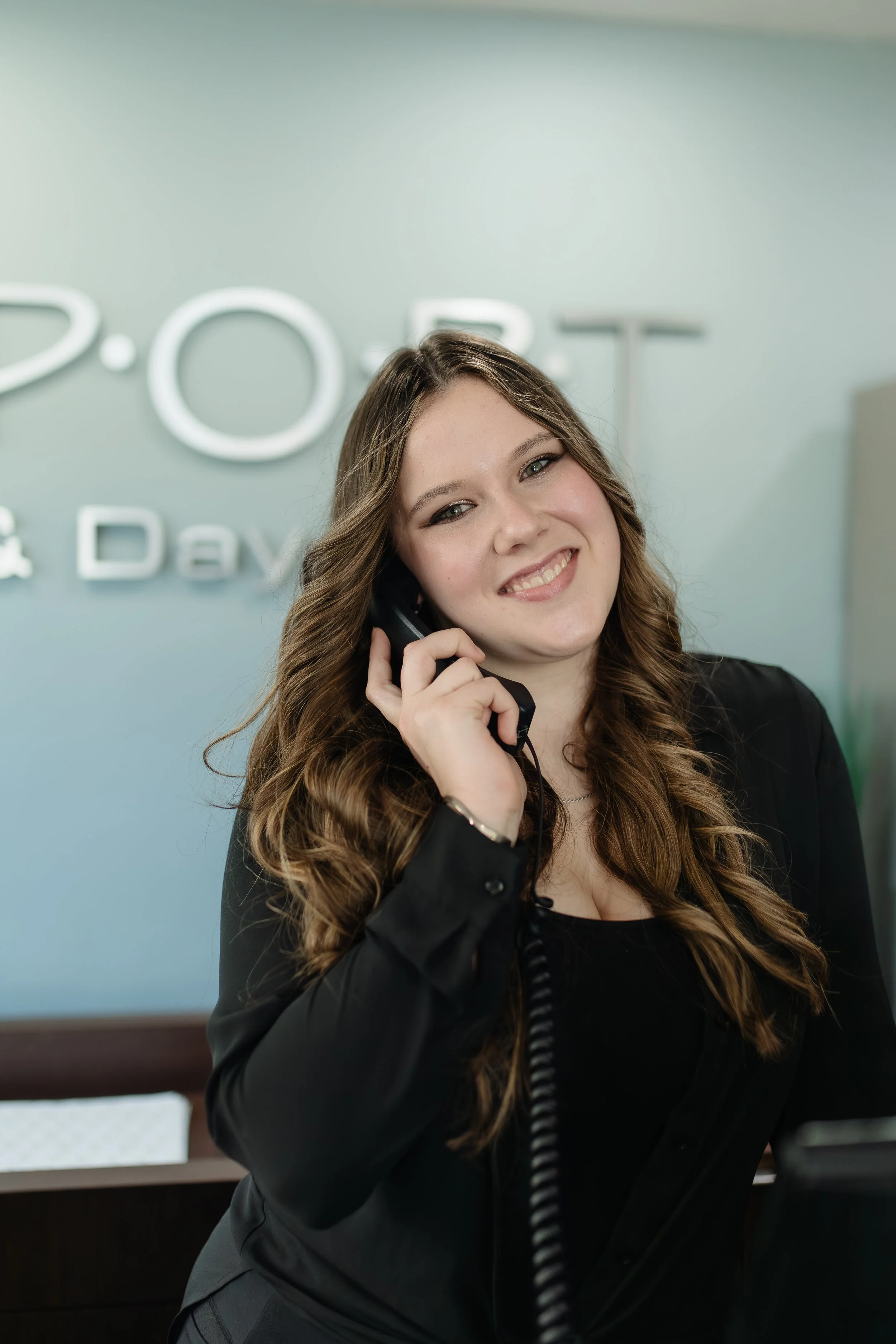 A young woman with long wavy brown hair smiling while holding a black landline phone to her ear, standing in front of a sign that reads 'P.O.S.T.' on the wall.