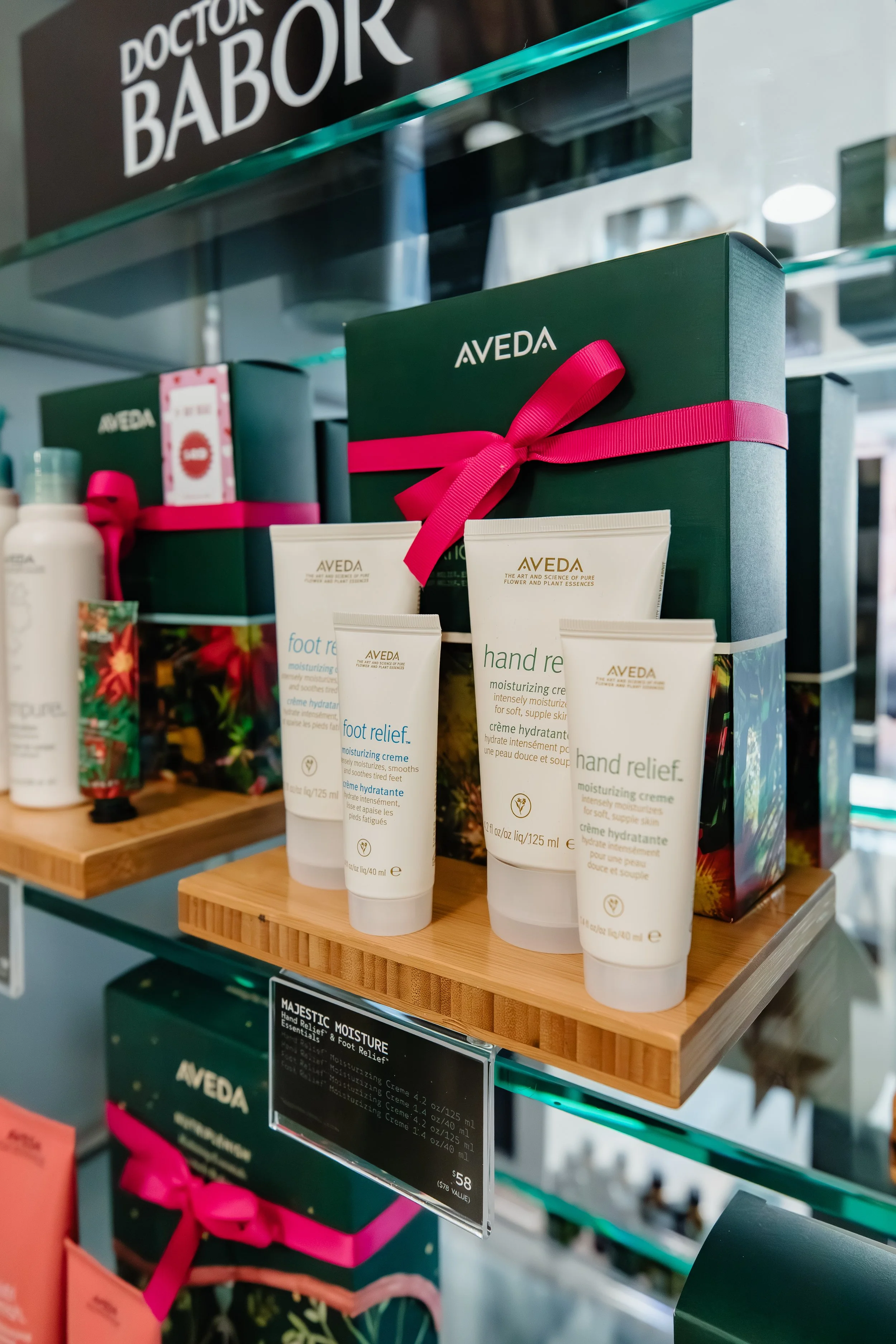 A display of Aveda skincare products, including hand relief and foot relief moisturizing creams, in gift boxes with pink ribbons on glass shelves.