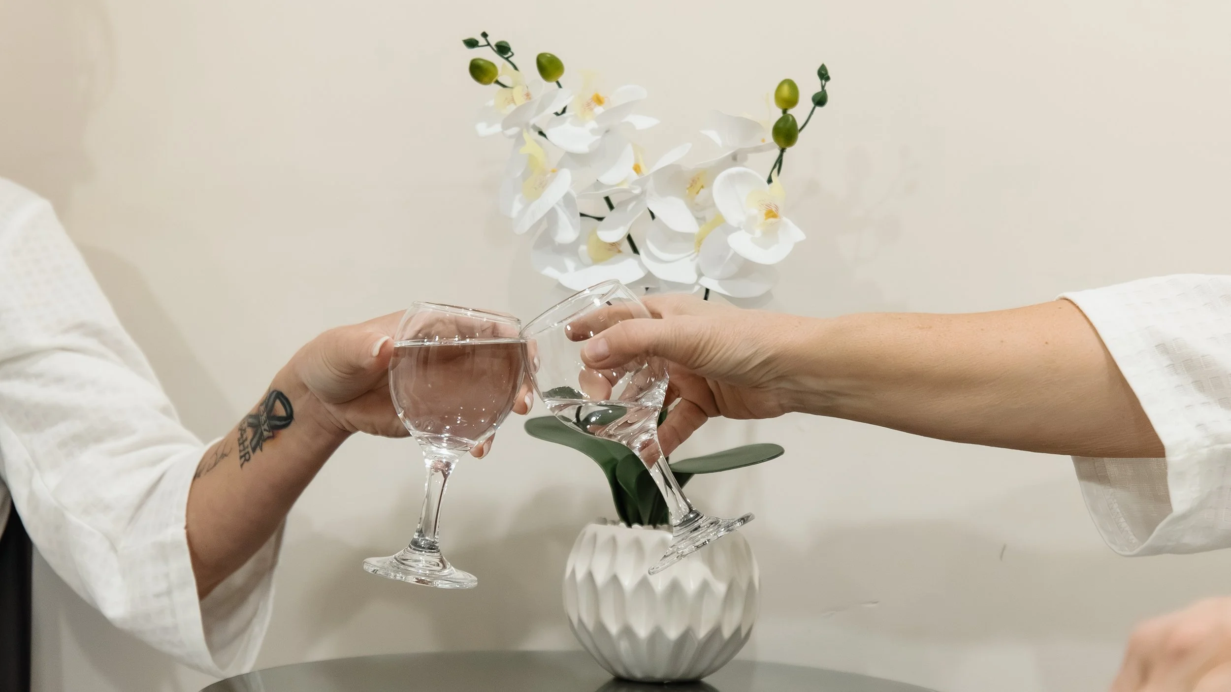 Two people clinking glasses of water in front of a white orchid plant in a decorative vase.