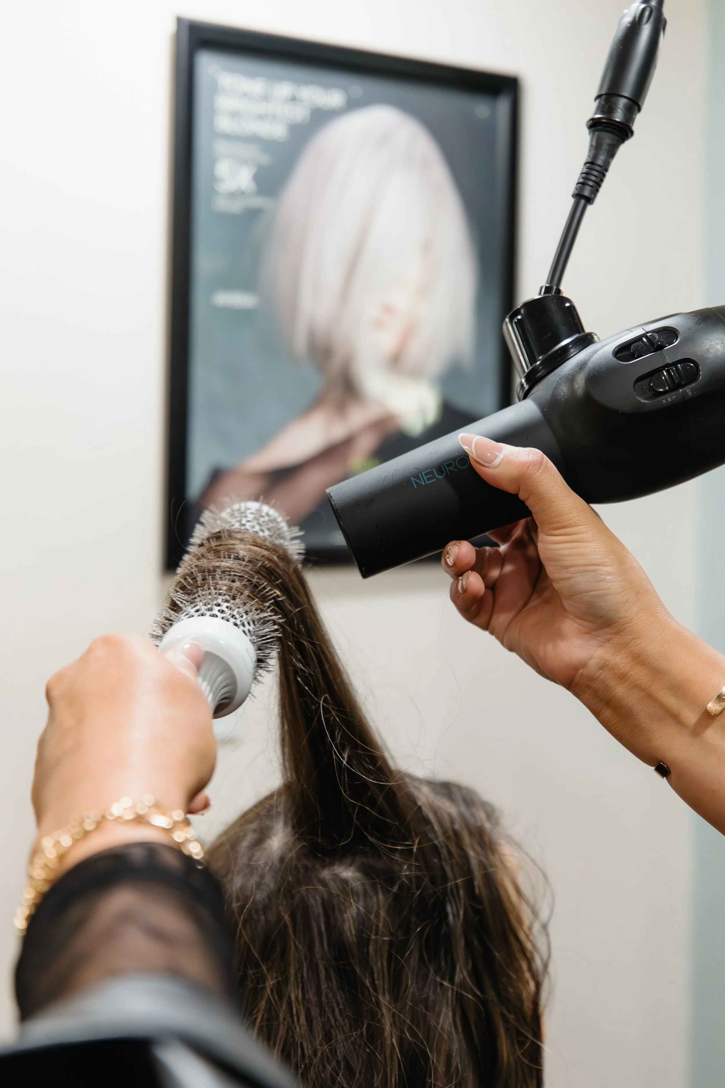 A person getting a hair coloring treatment, with a brush in one hand and a spray device labeled 'NEURO' in the other, in front of a framed hair color chart on the wall.