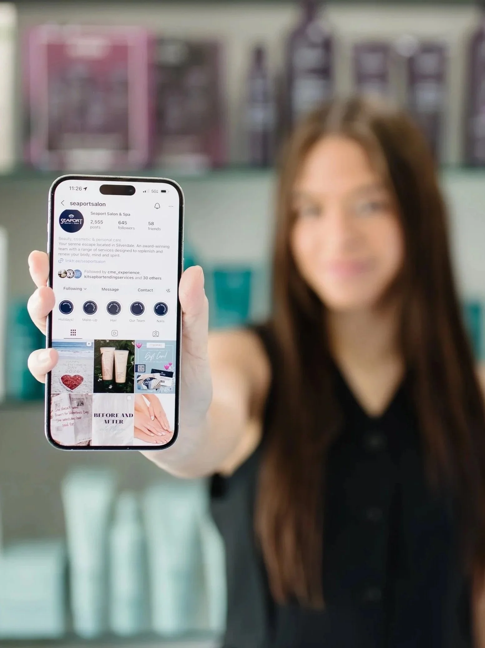 A woman holding a smartphone displaying the Instagram profile of Seaport Salon & Spa in a salon setting.