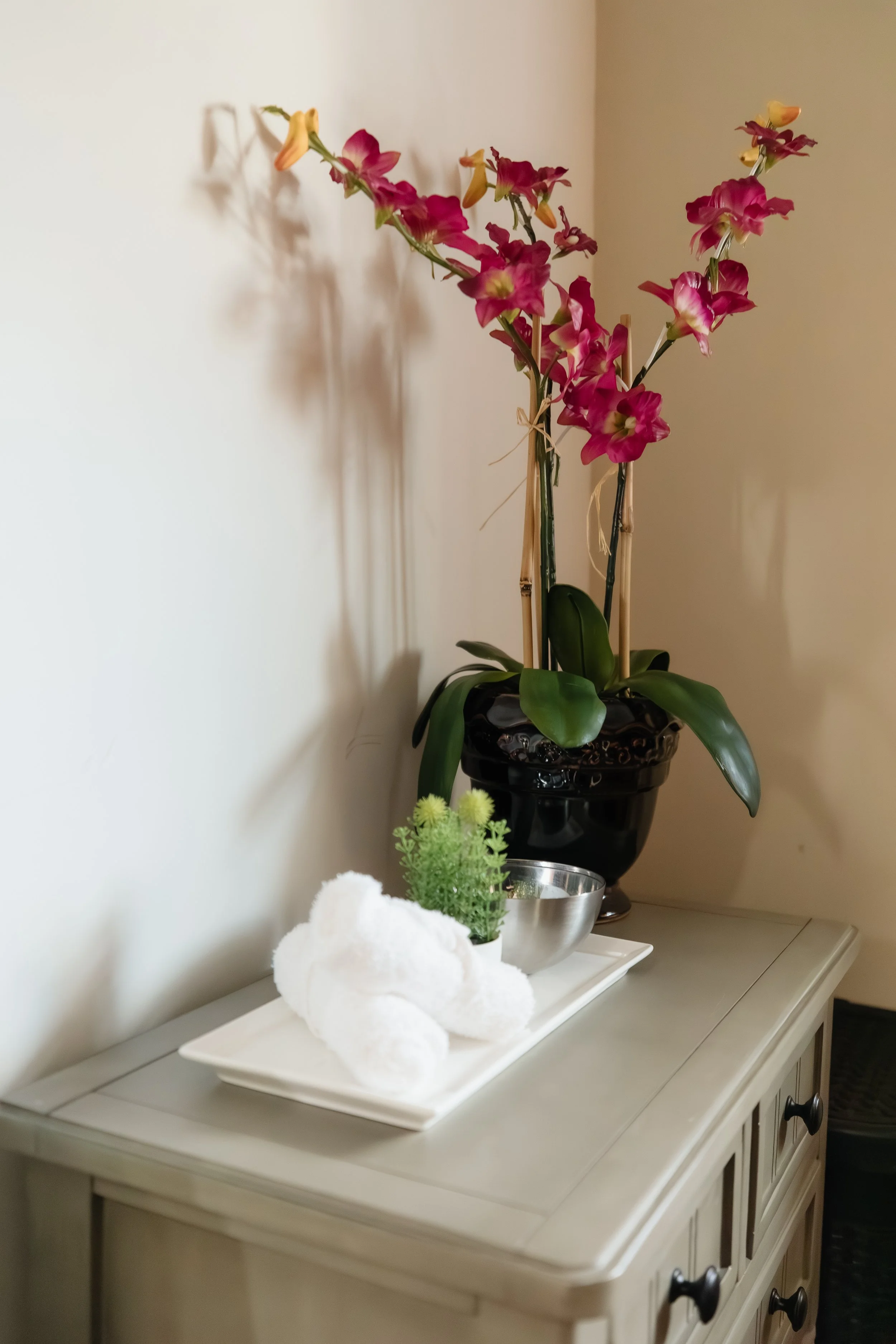 A white dresser with a black potted orchid, a towel folded on a tray, a small potted plant, and a silver bowl on top.