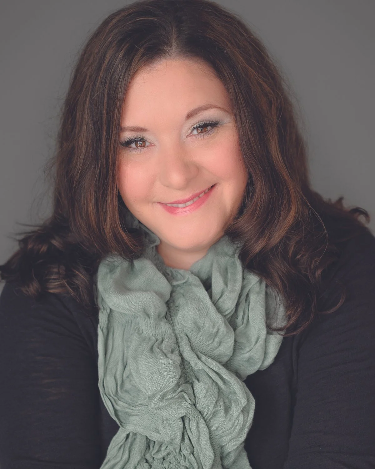 A woman with brown hair smiling, wearing a black top and a light green scarf against a gray background.
