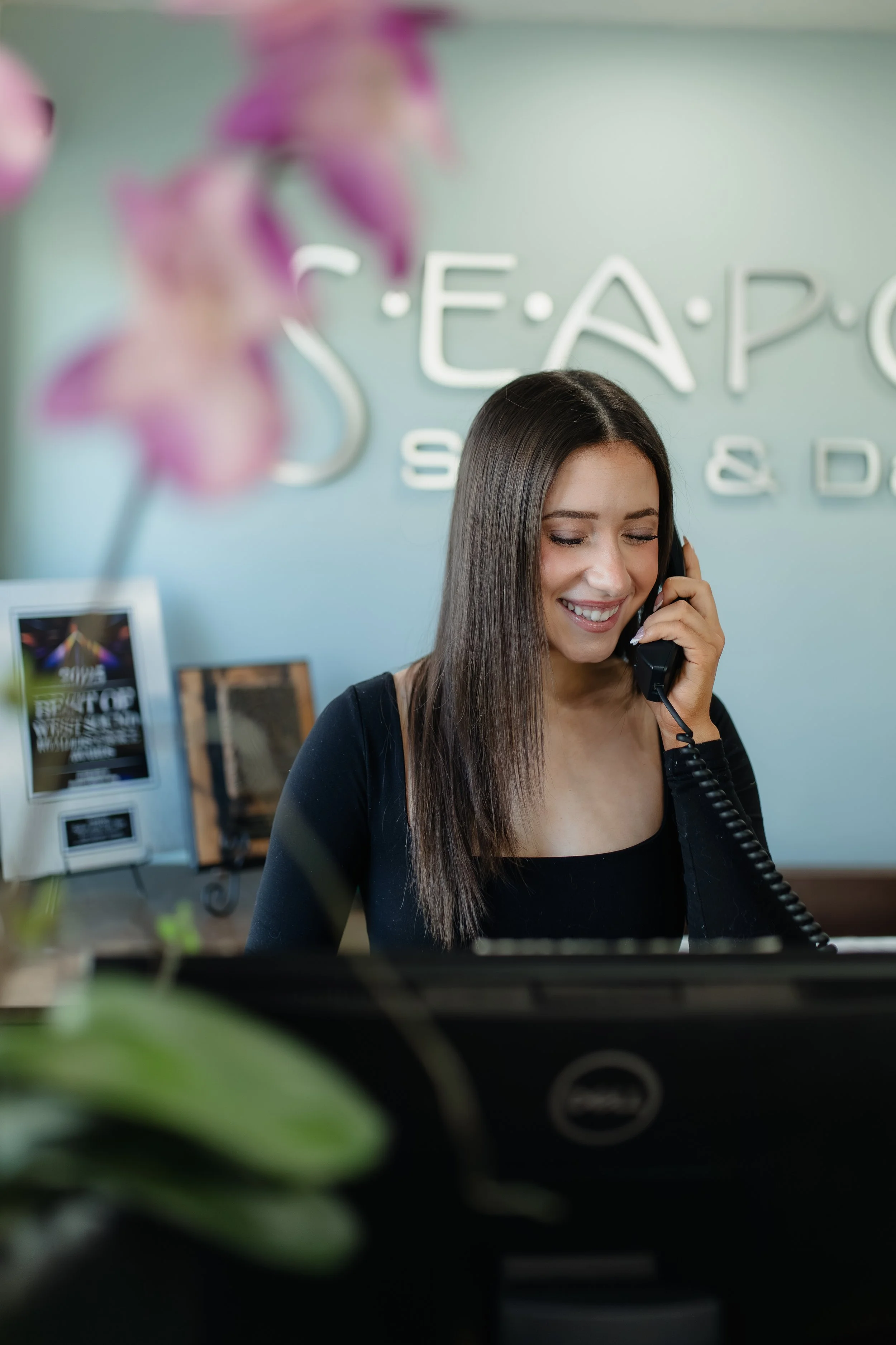 A smiling woman with long dark hair talking on a landline phone at a hotel reception desk.