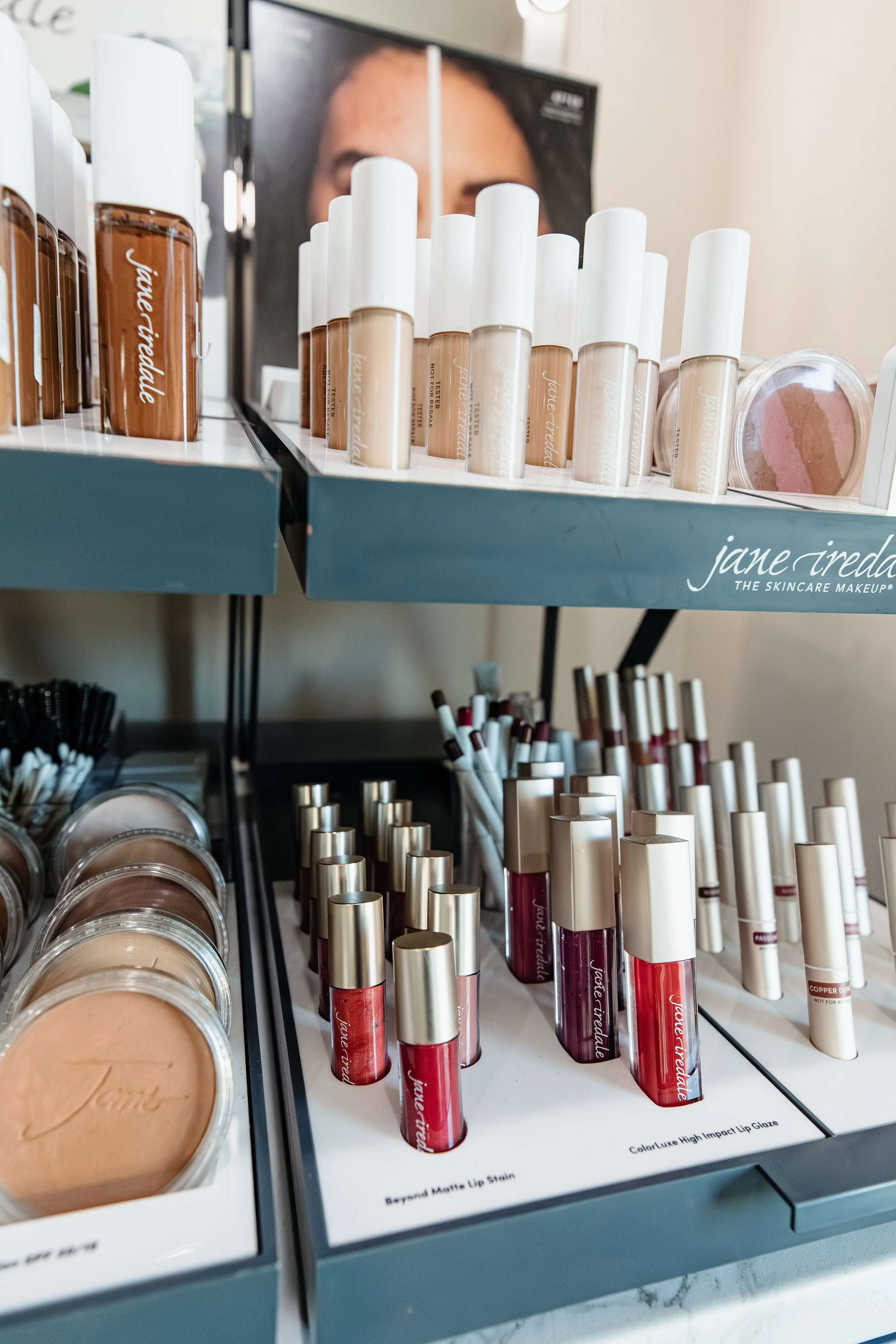 Display of Jane Iredale makeup products, including lip glosses, foundation bottles, and powders arranged on shelves.