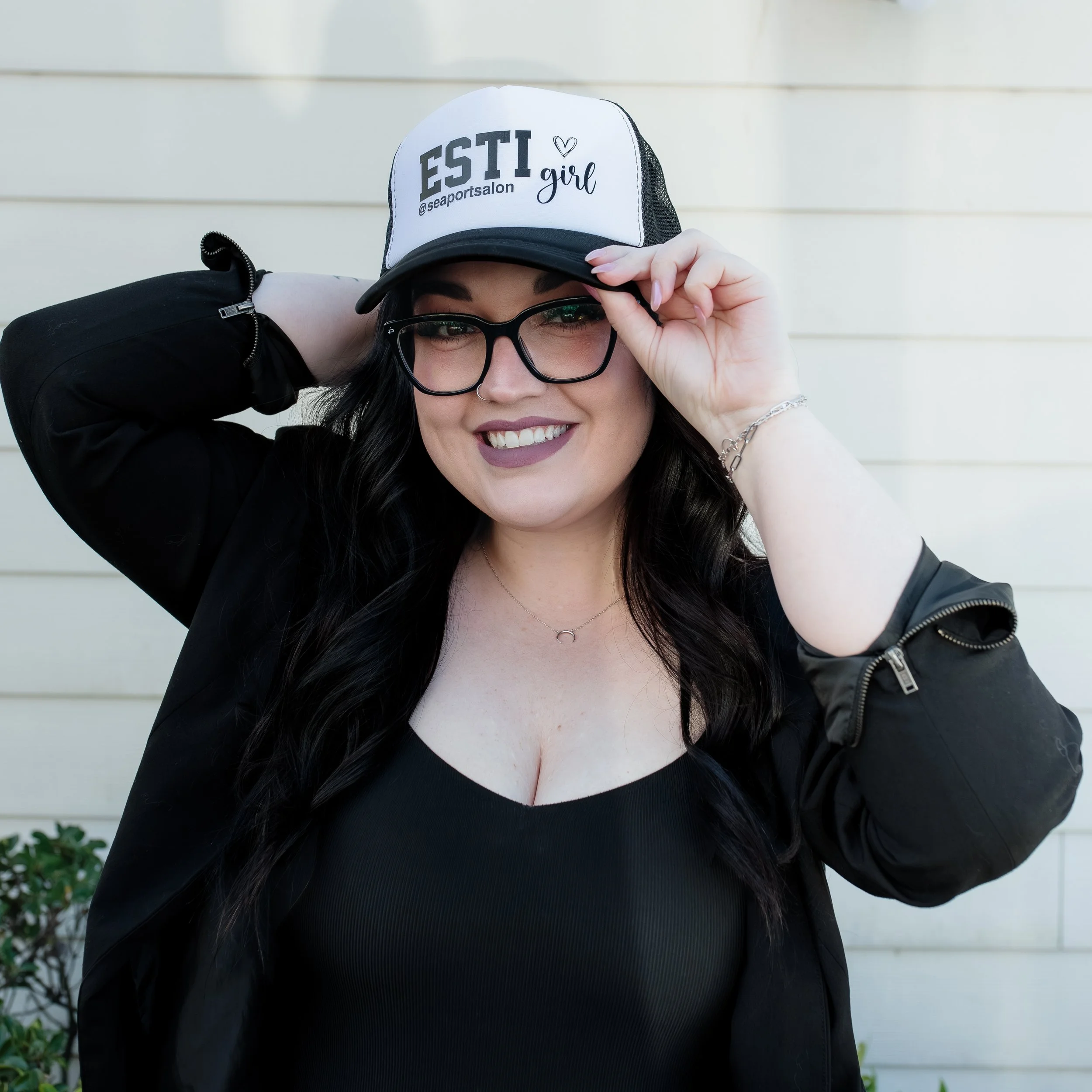 A woman with long dark hair, glasses, and a septum piercing, smiling, wearing a black top and jacket, adjusting a white and black trucker hat that says 'ESTI girl' and includes a social media handle.