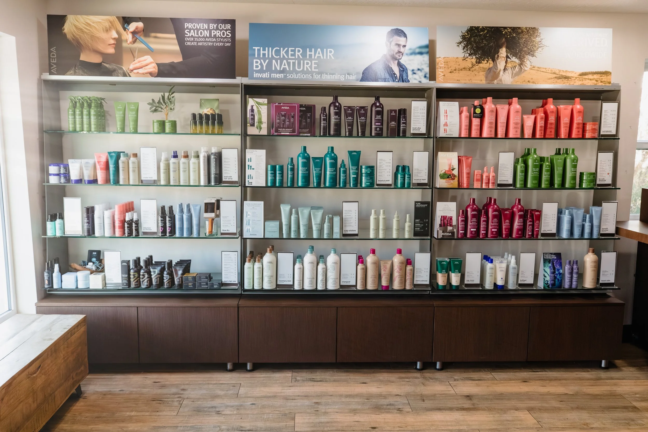 A wall display of hair care products organized by color, including shampoos, conditioners, and styling treatments, with promotional banners above highlighting hair thickening solutions and salon services.
