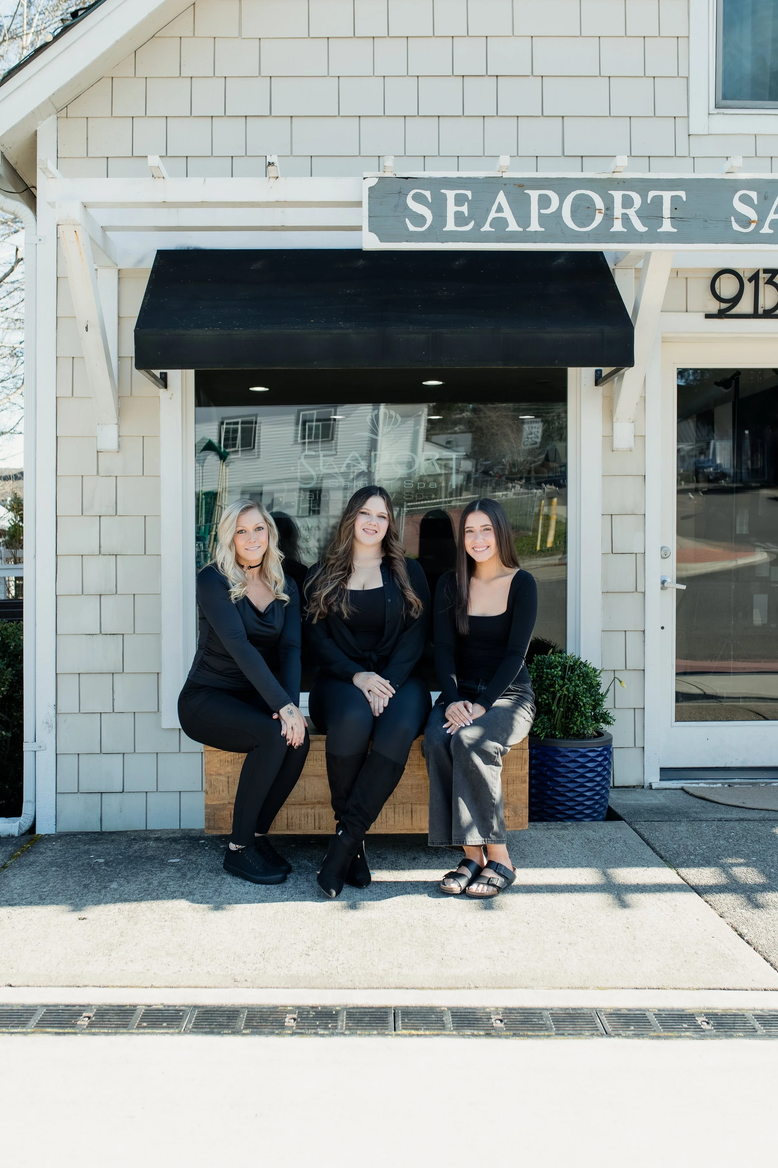 Three women sitting outside a spa called Seaport Spa, with a beige building and large window reflecting neighboring structures.