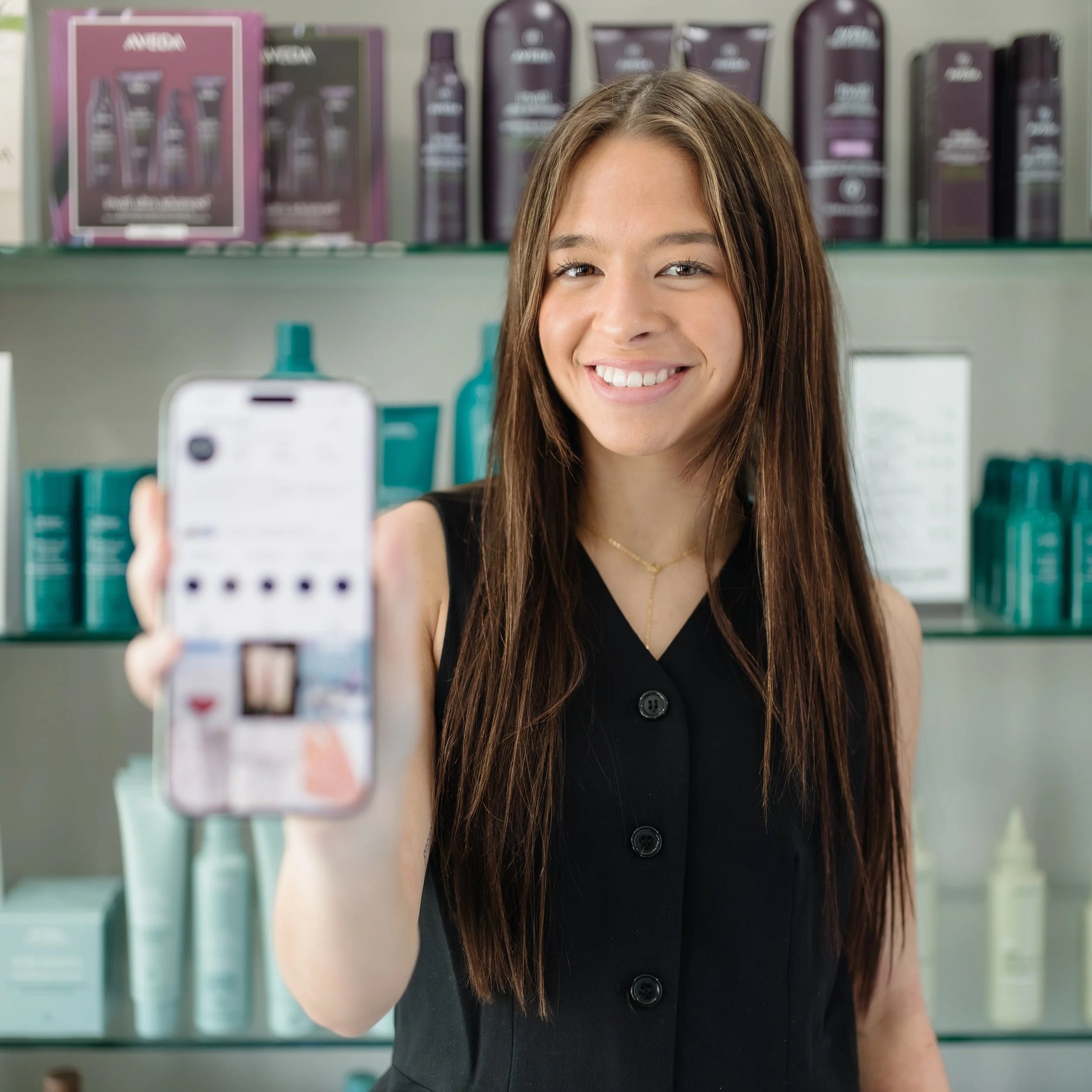 A young woman with long brown hair smiling as she holds a smartphone, taking a selfie in a store with shelves of hair care products in the background.