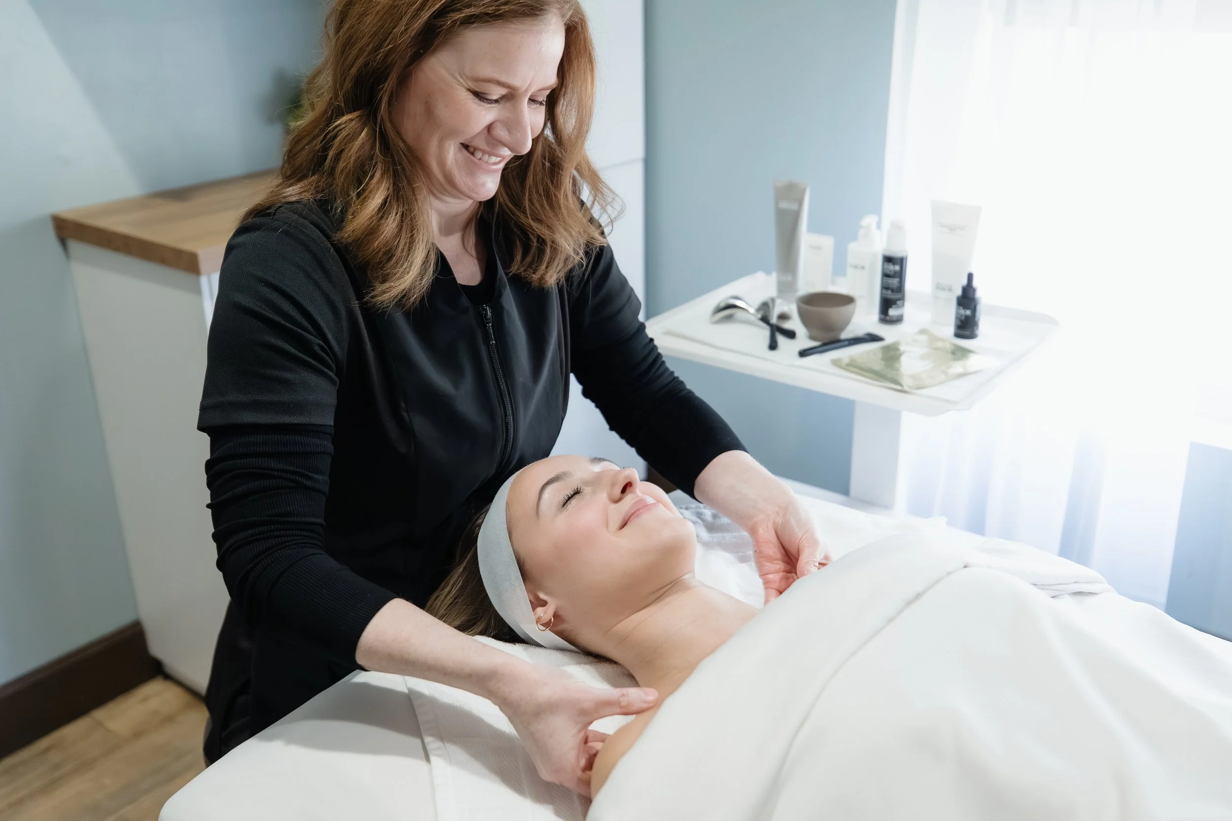 A woman receiving a facial treatment at a spa or clinic, lying on a treatment bed with her eyes closed, while a professional woman in black applies skincare.