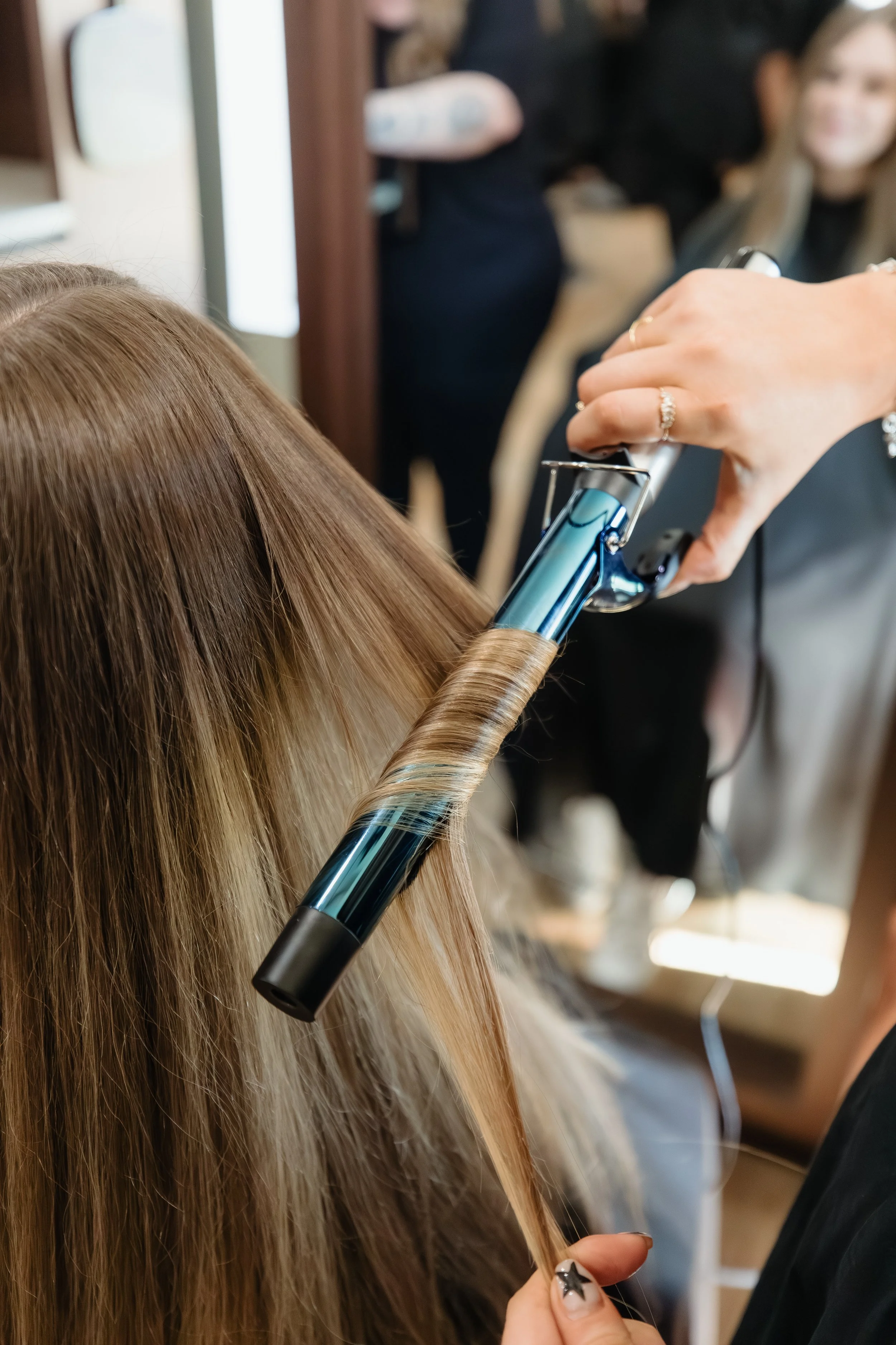 A person is curling a woman's long, blonde hair with a curling iron at a salon.
