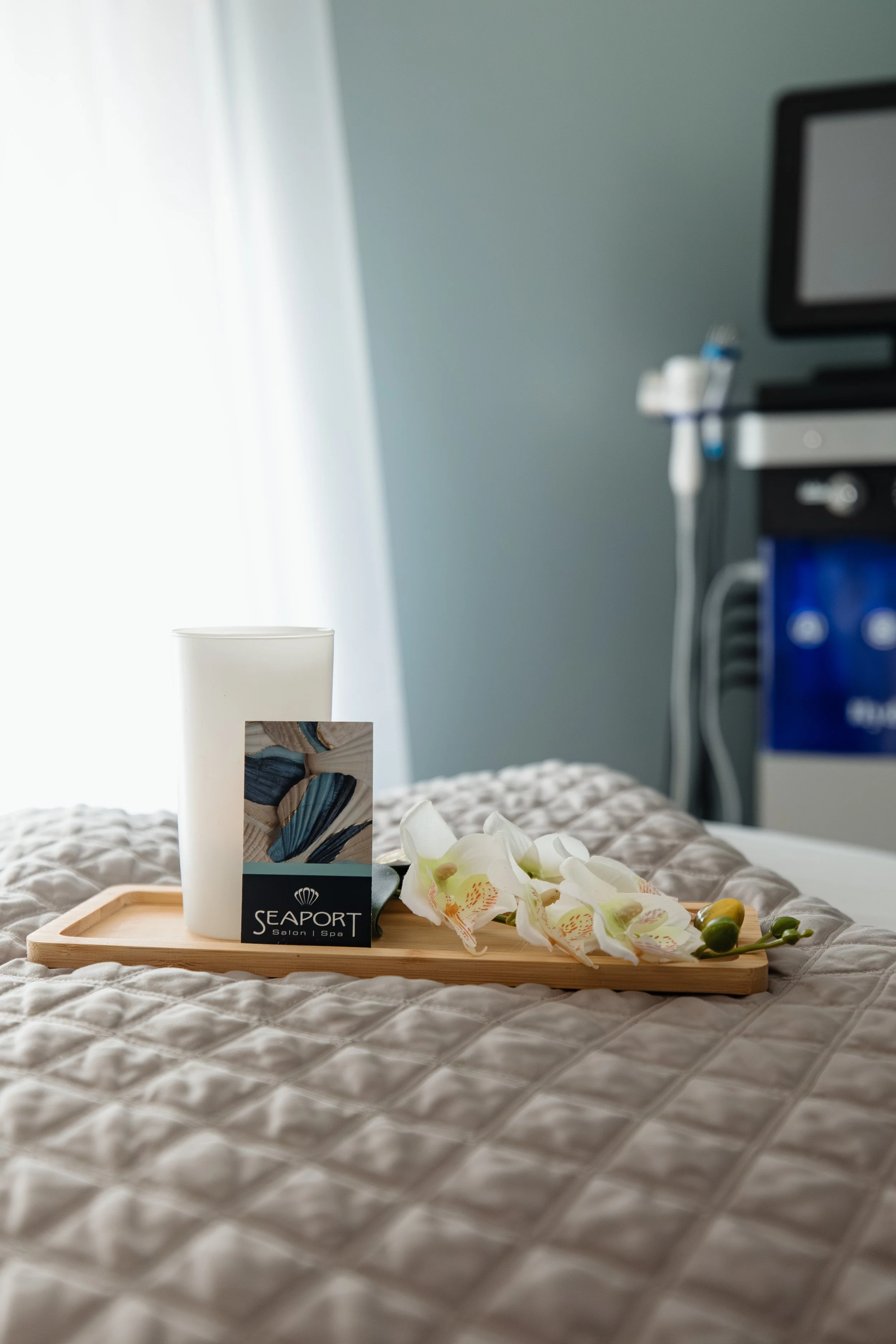 A hotel tray with a white cup, orchid flowers, and a card with 'Seaport Salon Spa' on it, placed on a bed with a quilted beige bedspread. In the background, there is medical equipment and a computer monitor in a room with gray walls and curtains.