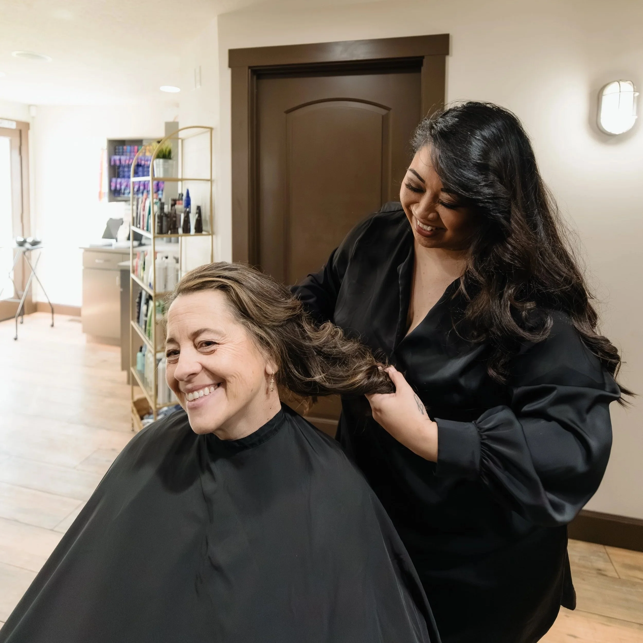 A woman with shoulder-length gray hair getting her hair styled by a smiling hairstylist with long black hair in a salon.
