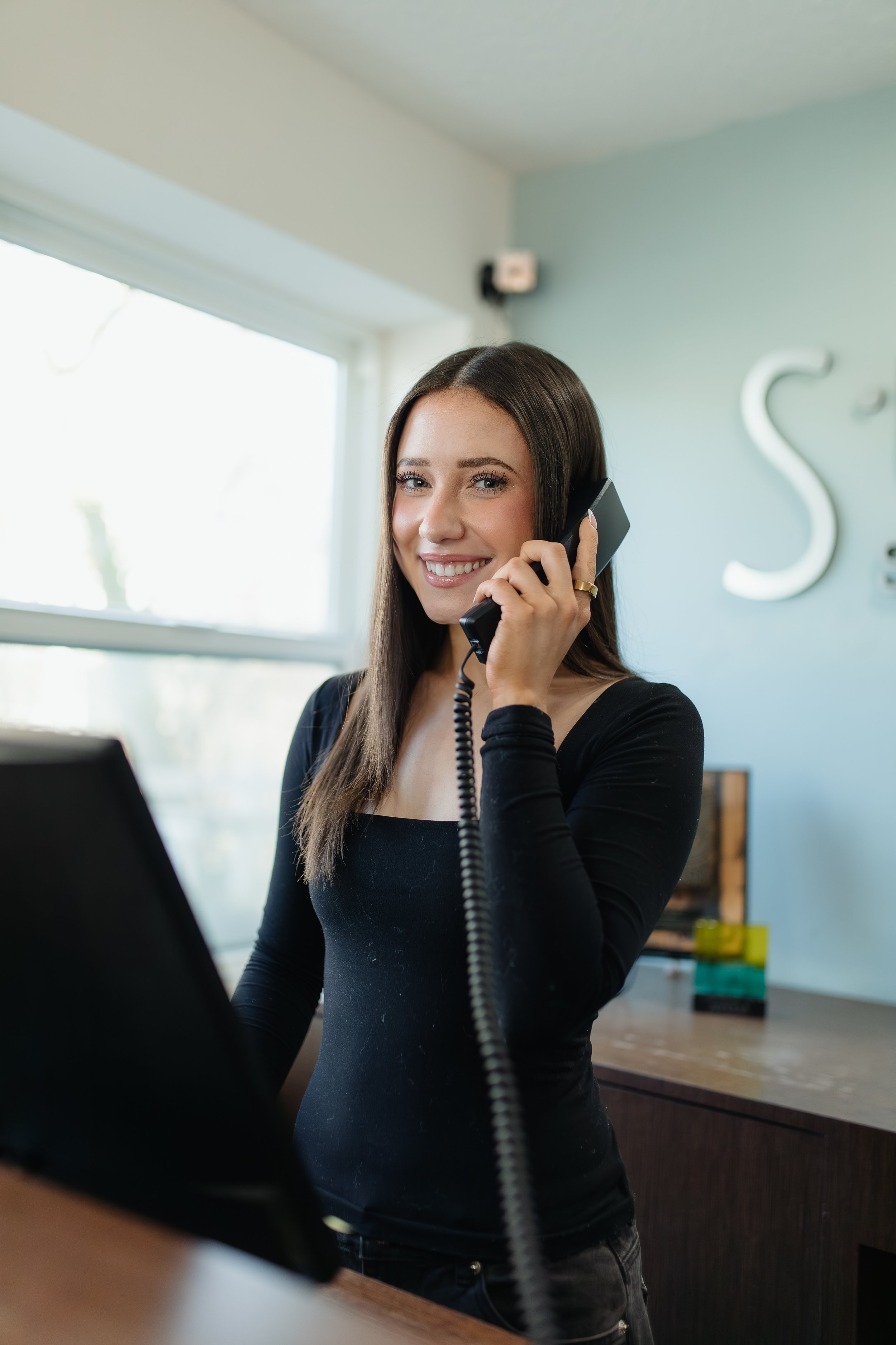 A woman with long brown hair and a black top smiling while talking on a landline phone.