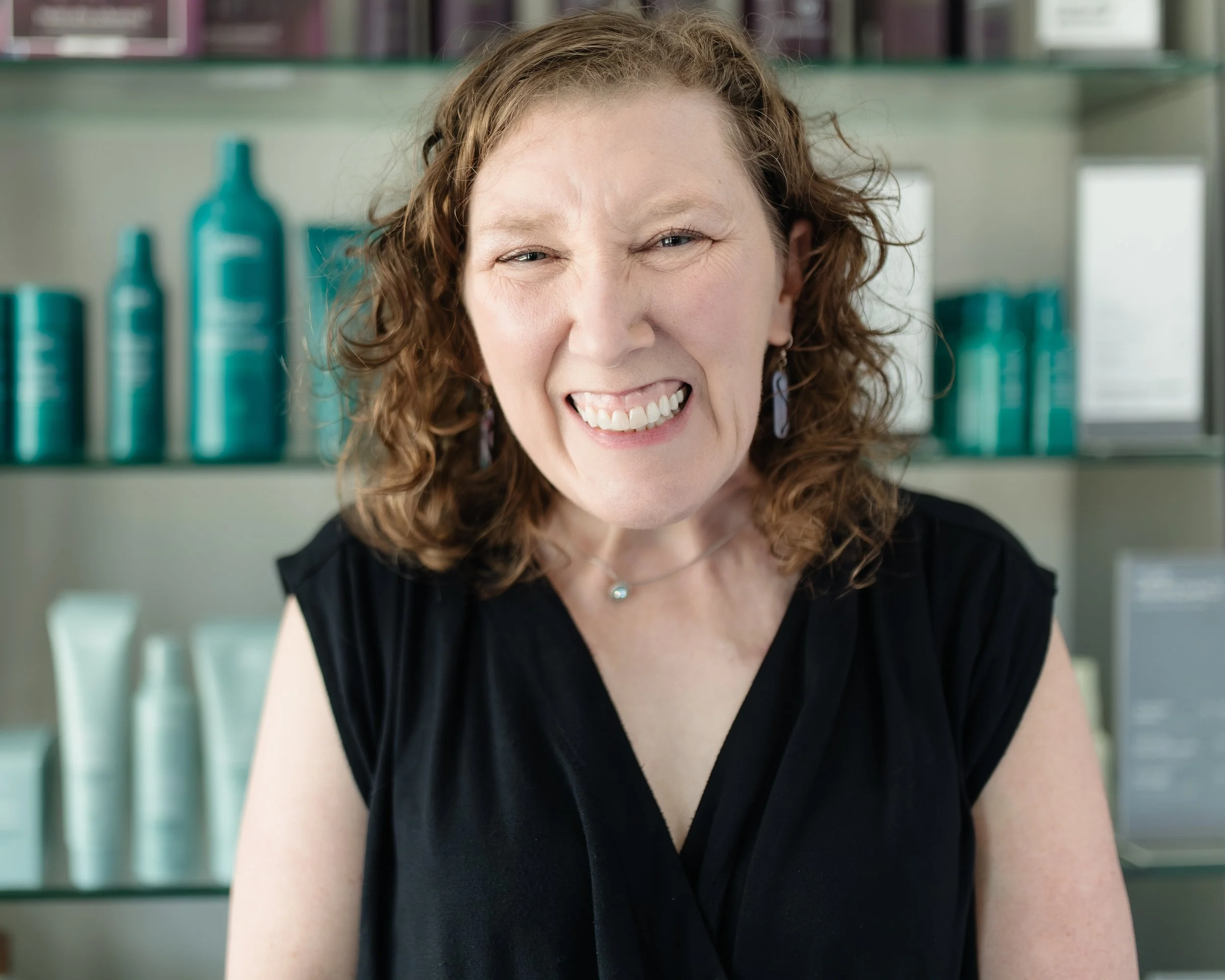 A woman with curly brown hair smiling and showing her teeth at the camera, wearing a black top, earrings, and a necklace, standing in front of shelves with hair care products.