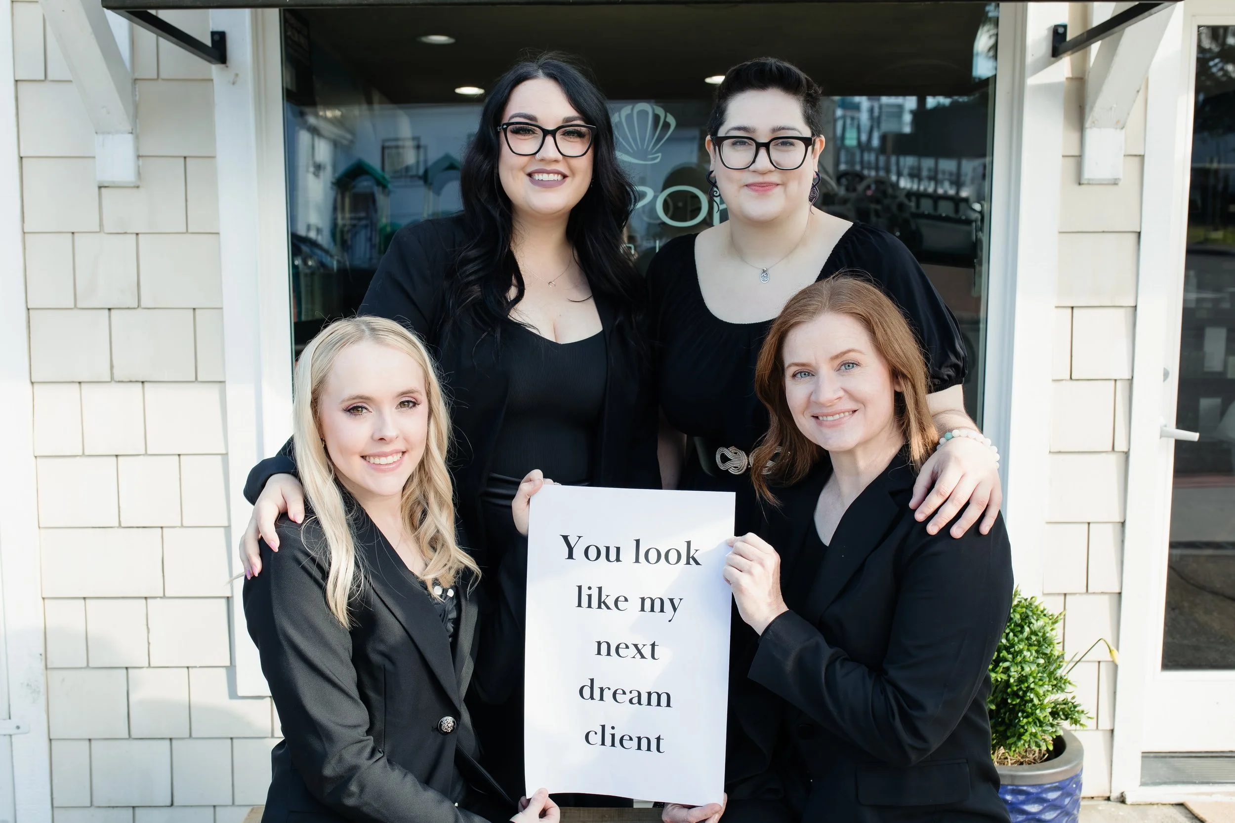 Four women dressed in black, standing and sitting outside a building, holding a sign that reads 'You look like my next dream client'.