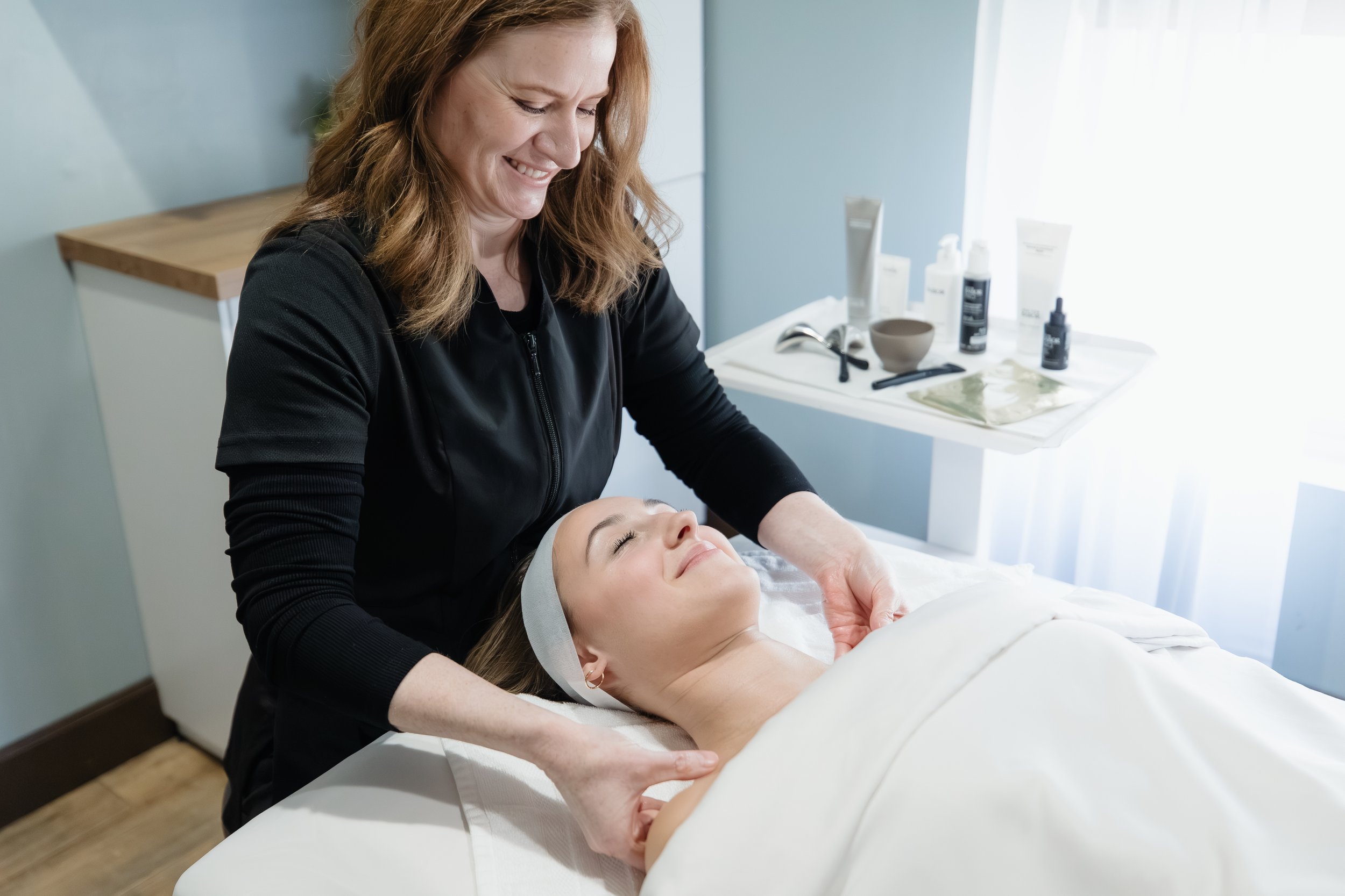 A woman lying on a treatment bed with her eyes closed, receiving a facial from a skincare professional in a spa or clinic setting.