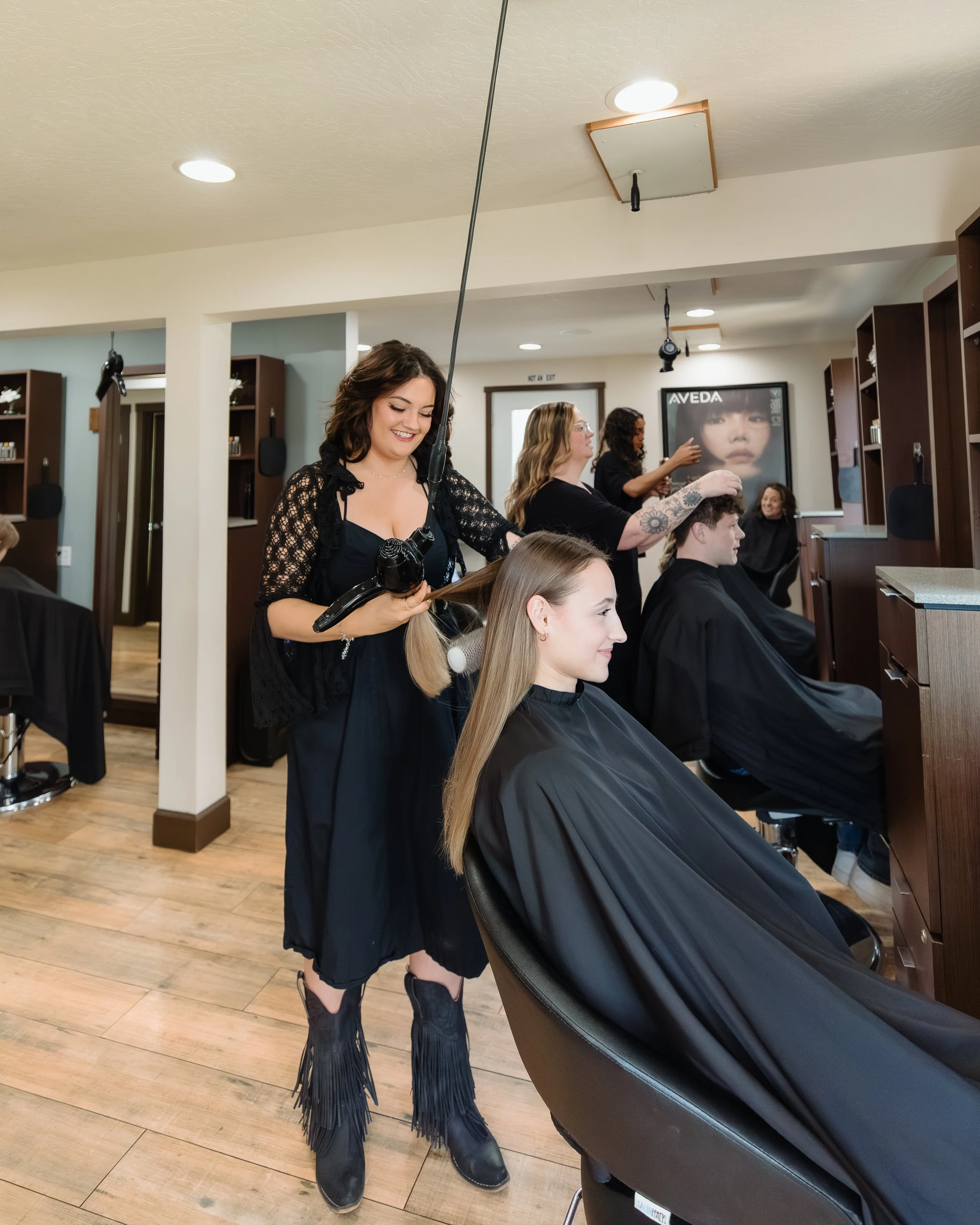 A hairstylist drying a woman's hair in a salon, with other clients and stylists visible in the background.