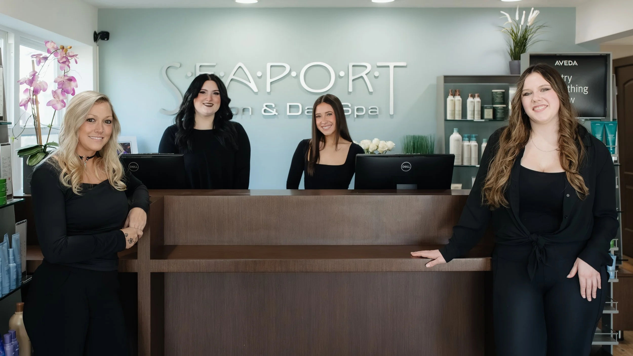 Four women at the reception desk of a spa and massage salon, smiling. The salon has a modern, clean look with shelves of products and a sign that reads 'SEAPORT Spa & Day Spa' in the background.
