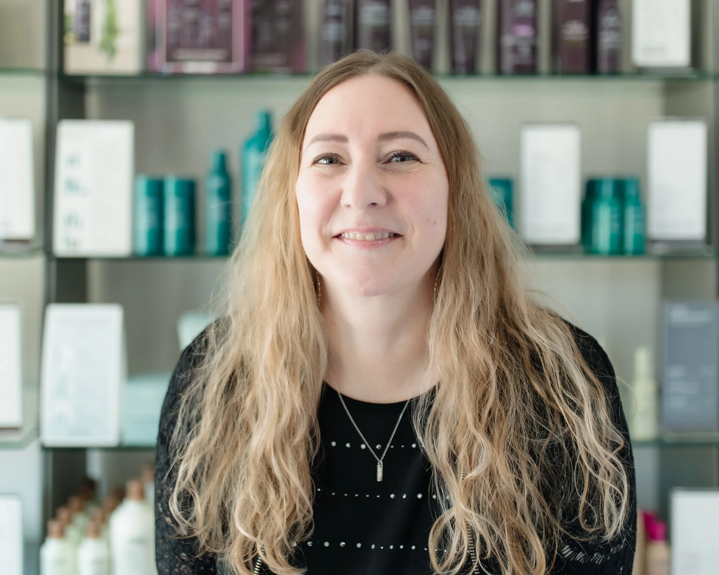 A woman with long, curly blonde hair smiling in front of store shelves with hair care products.